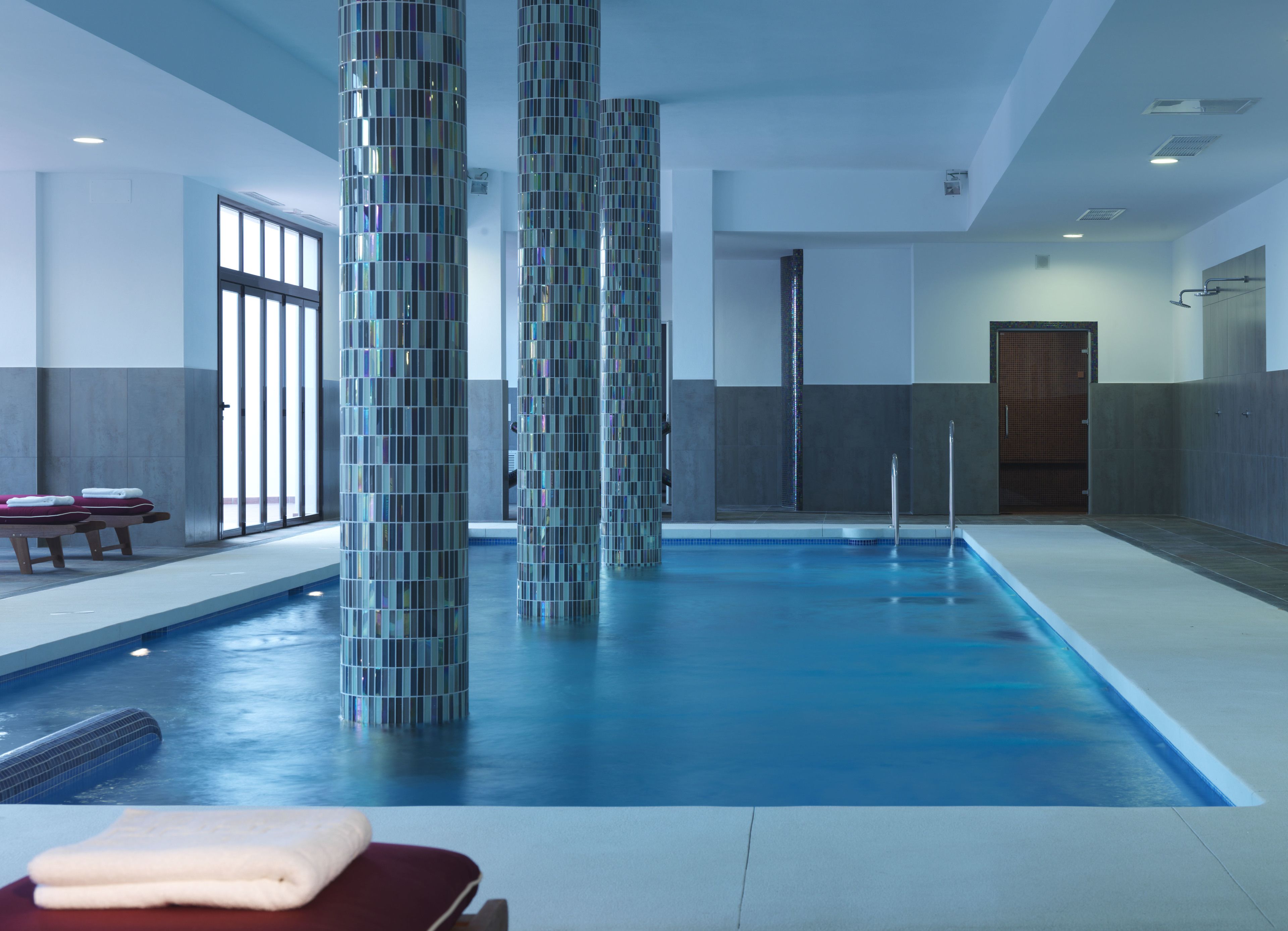 A modern indoor swimming pool with clear glass pillars, a lounge area with benches, and a door leading to another room.