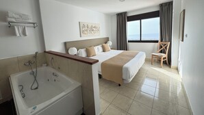 Habitación de hotel con una cama grande, una bañera, una silla y vistas a la ciudad a través del ventanal.