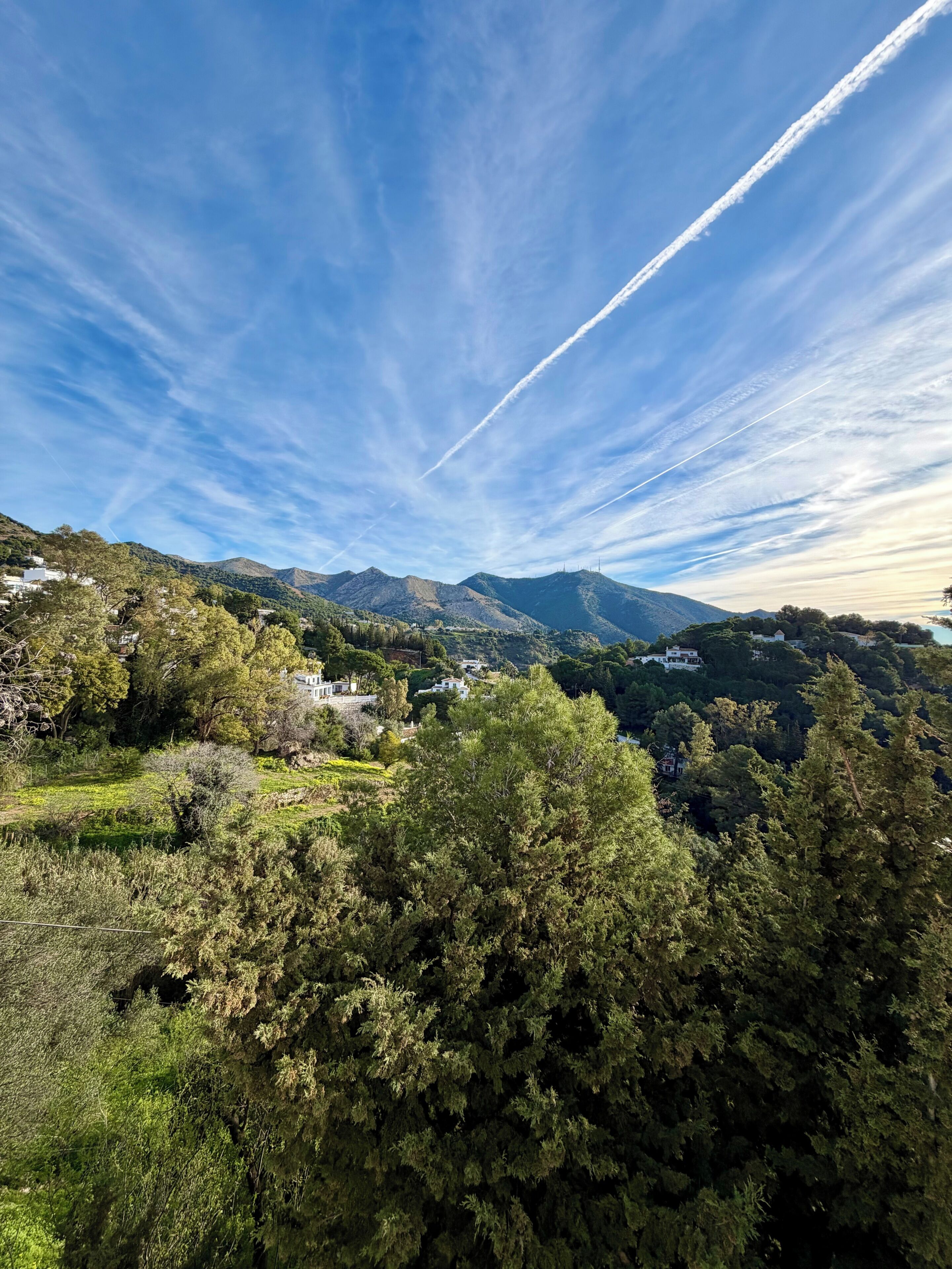 Un paisaje montañoso con edificios dispersos, vegetación densa y cielo despejado con estelas de condensación.