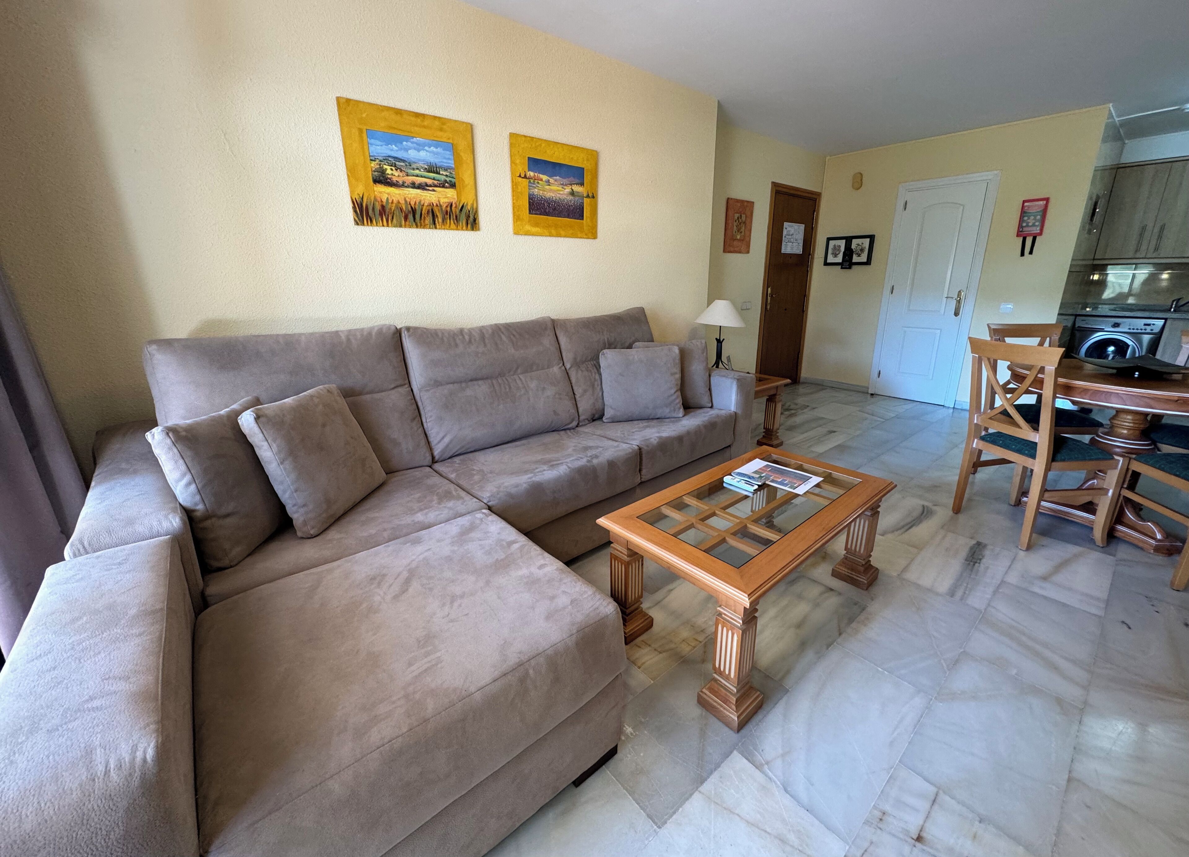 Una sala de estar con un sofá seccional color beige, una mesa de centro con cubierta de vidrio y un comedor con sillas de madera y una mesa.