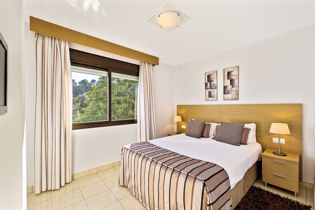 A hotel room with a large bed, a nightstand, a lamp, and a view of greenery outside.