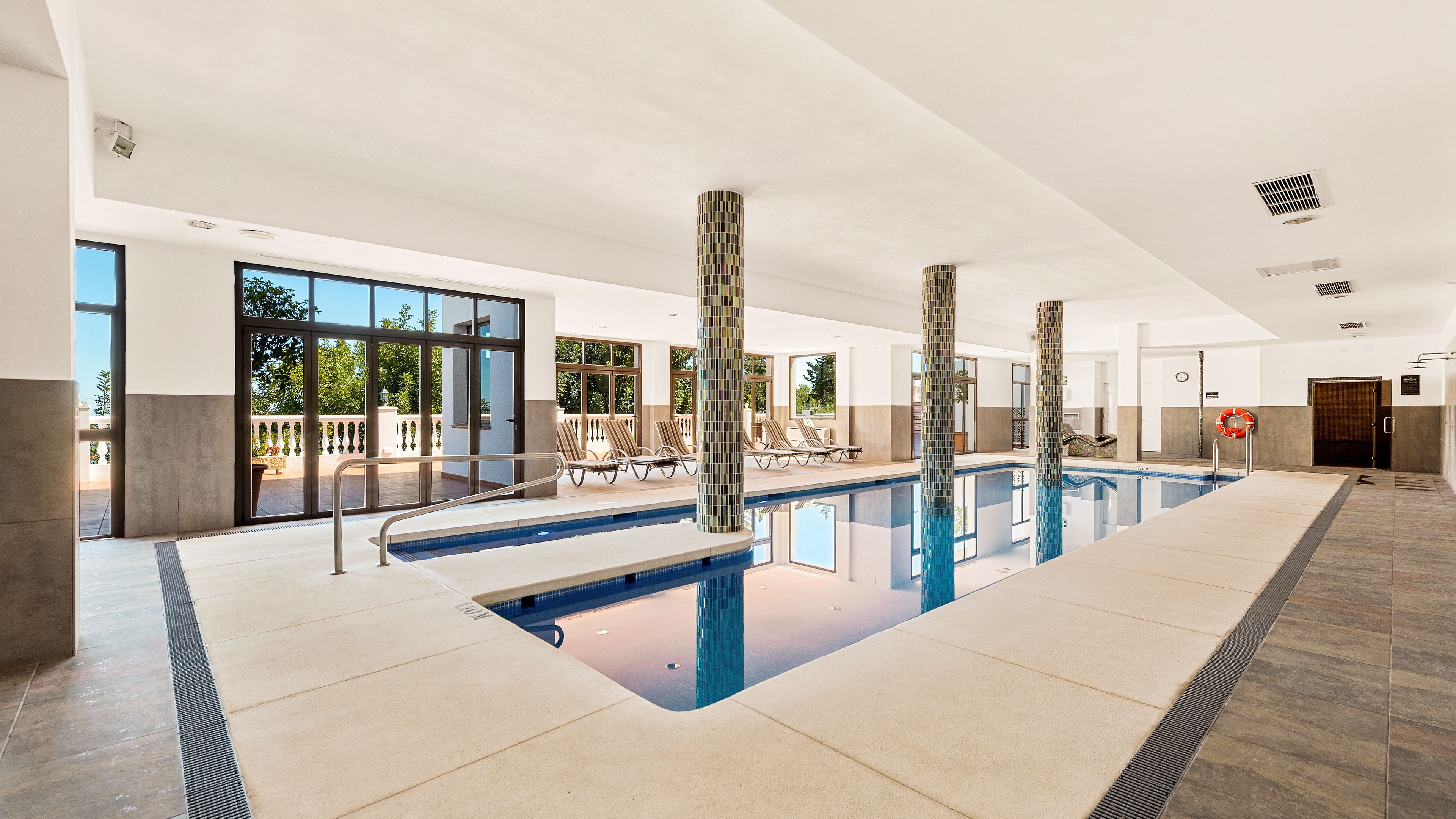 A modern indoor swimming pool with clear glass walls, sun loungers, and tiled columns.