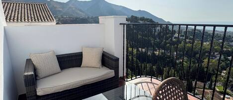 A balcony with a wicker sofa and a glass table, overlooking a mountainous landscape.
