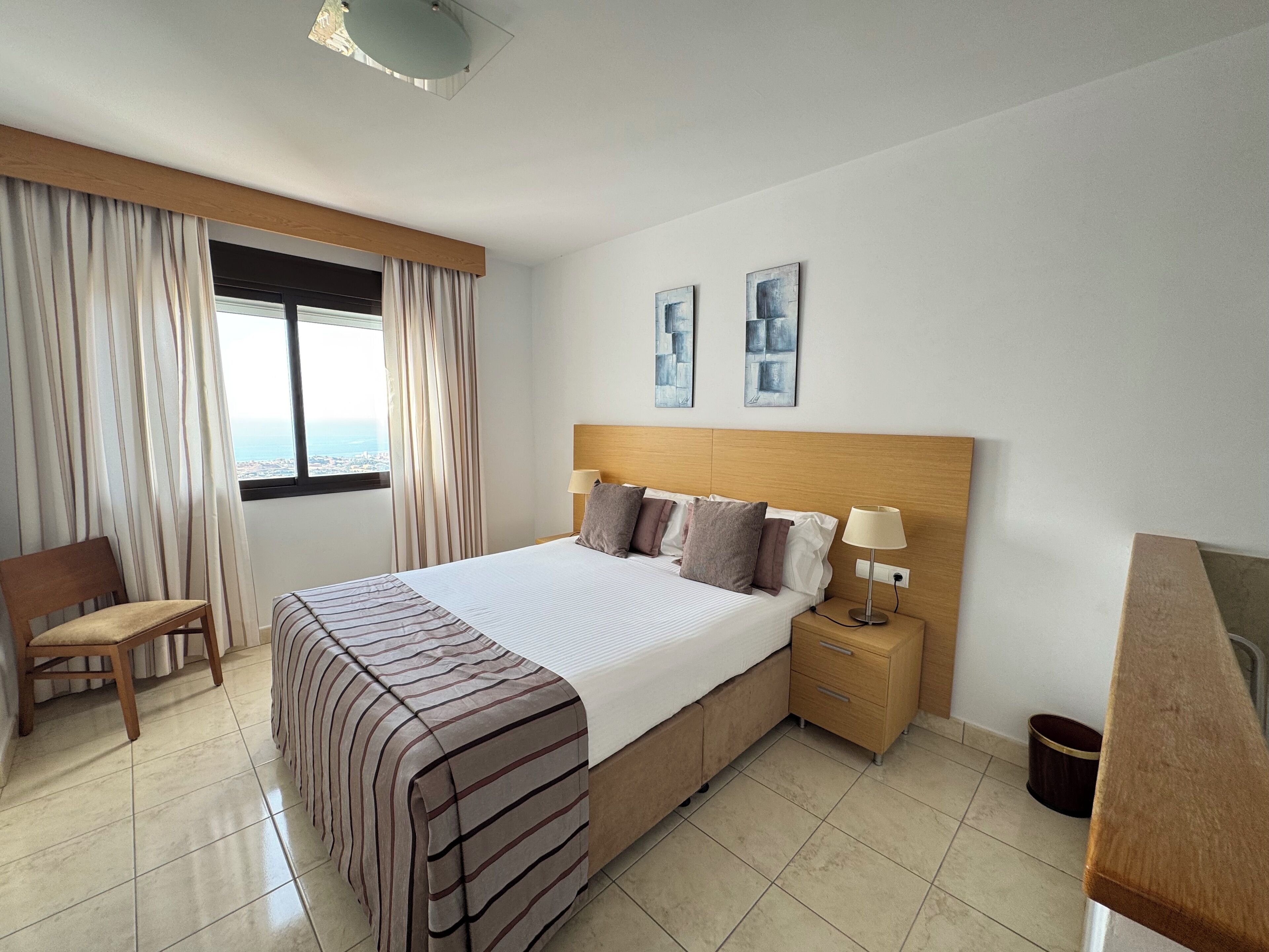 Habitación de hotel con una cama grande, cabecera de madera, mesita de noche con lámpara, una silla y vista a la ciudad desde la ventana.