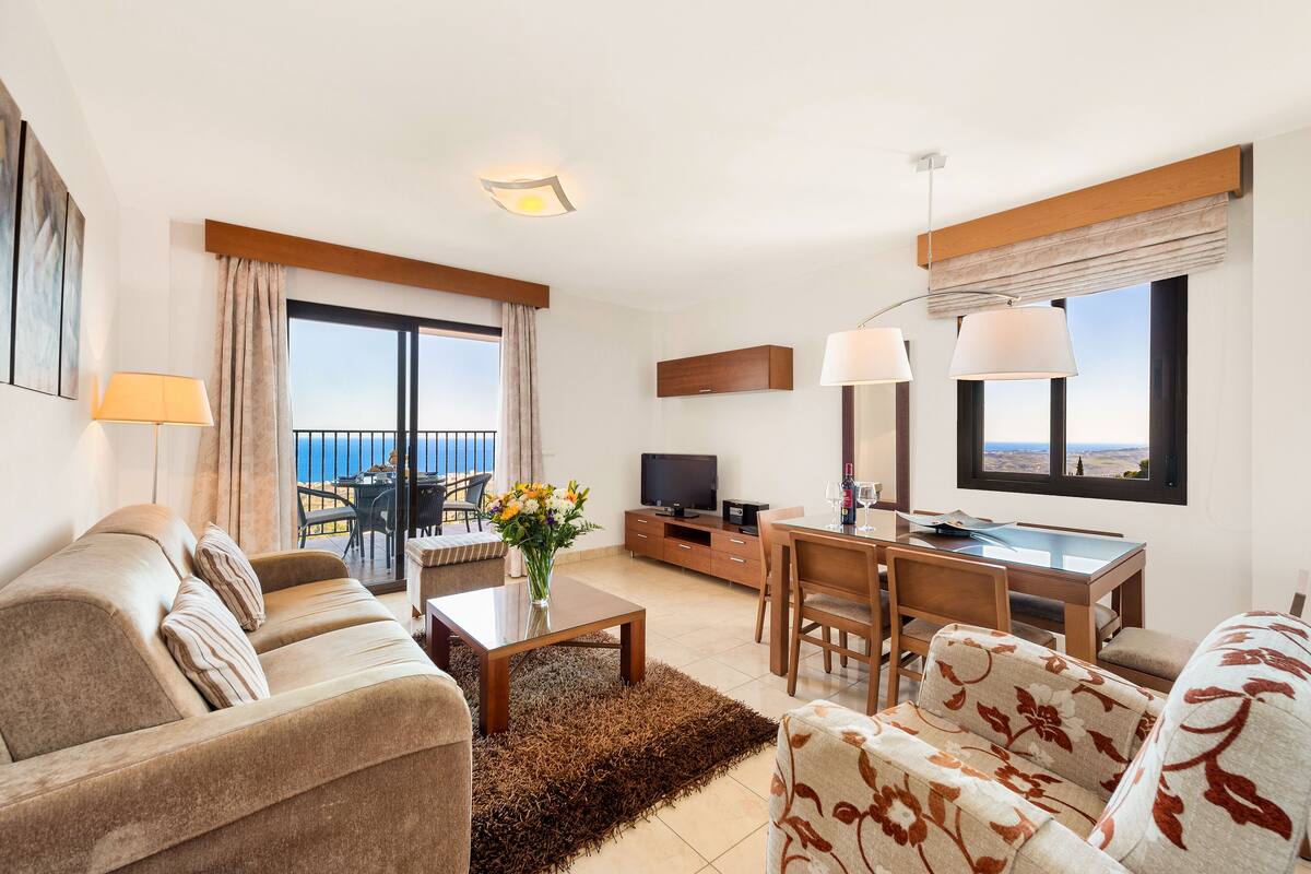 A modern hotel room with a sofa, a dining table, a TV, and a balcony with a view of the sea.