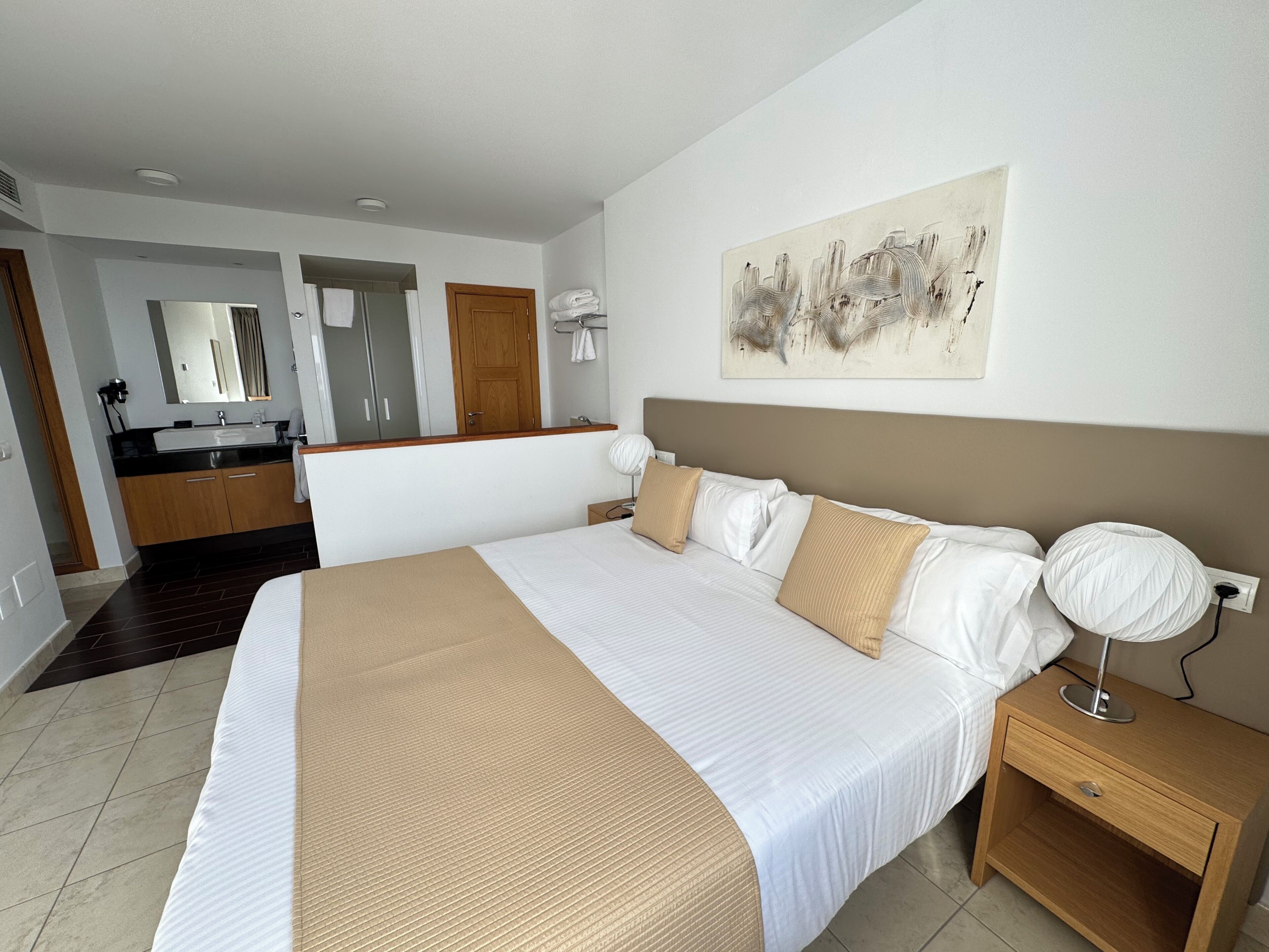 Una habitación de hotel con una cama grande, una mesita de noche de madera, una lámpara de noche, un baño con lavamanos y espejo, y un cuadro en la pared.