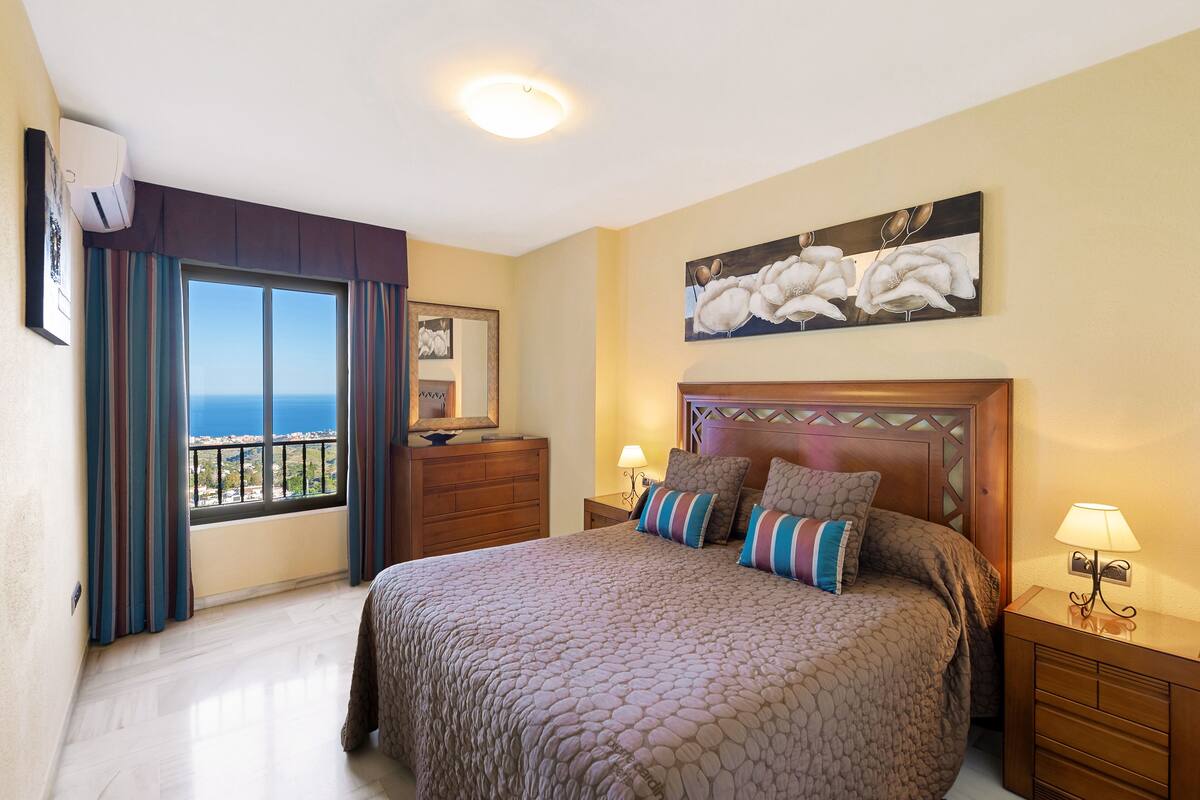 A bedroom with a large bed, a nightstand, a dresser, a window with curtains, and a painting on the wall.