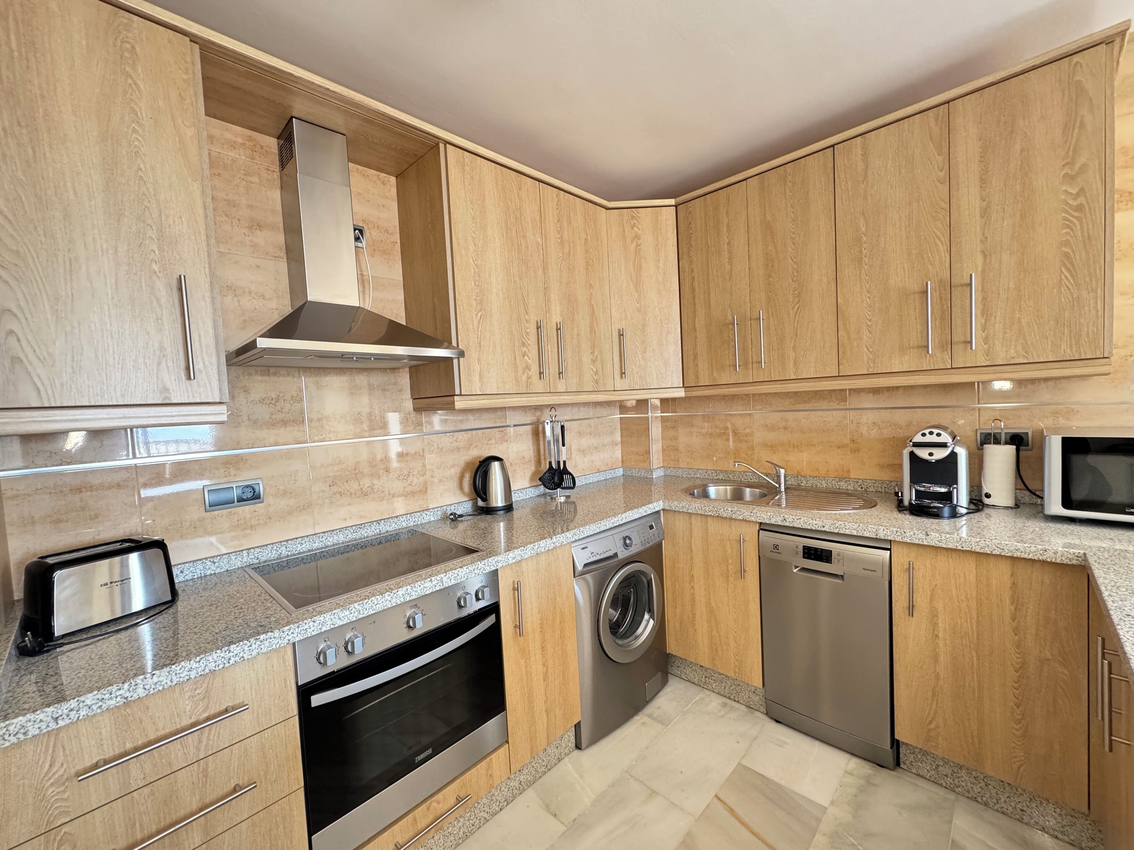 A modern kitchen with wooden cabinets, stainless steel appliances, a built-in oven, a washing machine, and a dishwasher.