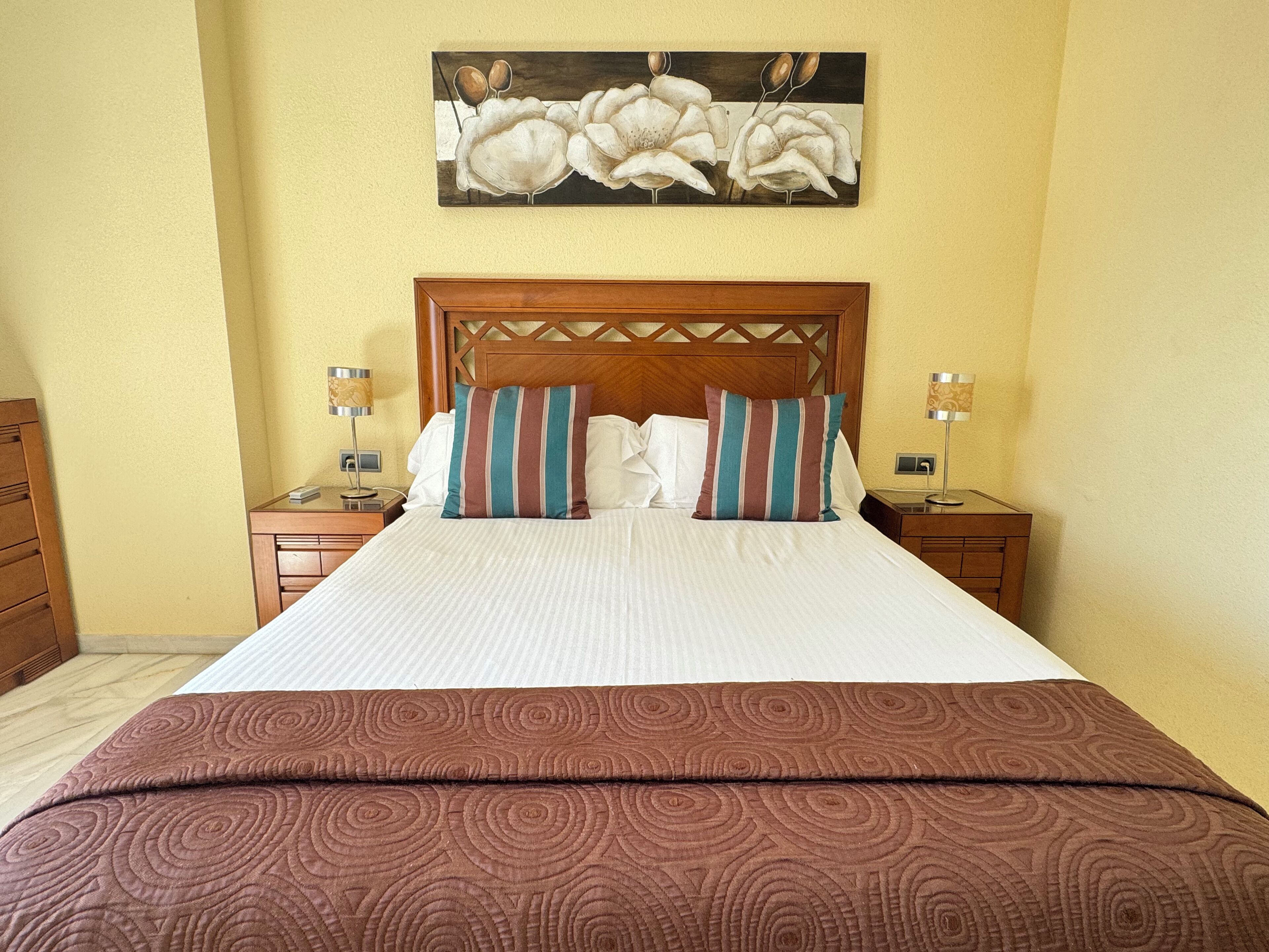 Una habitación de hotel con una cama grande, dos mesitas de noche, un cabecero de madera y un cuadro de flores en la pared.