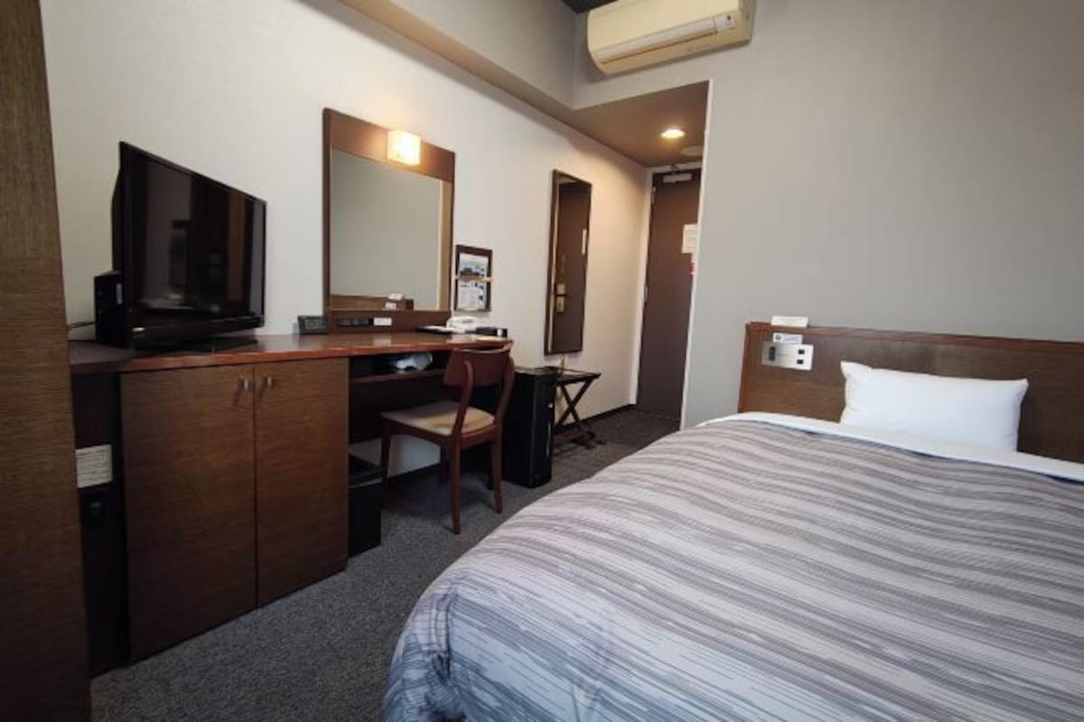Una habitación de hotel con cama, escritorio, silla, televisor y espejo.
