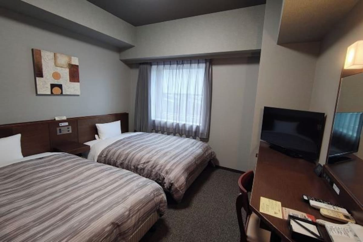 Una habitación de hotel con dos camas, un escritorio con televisor y una ventana con cortinas.