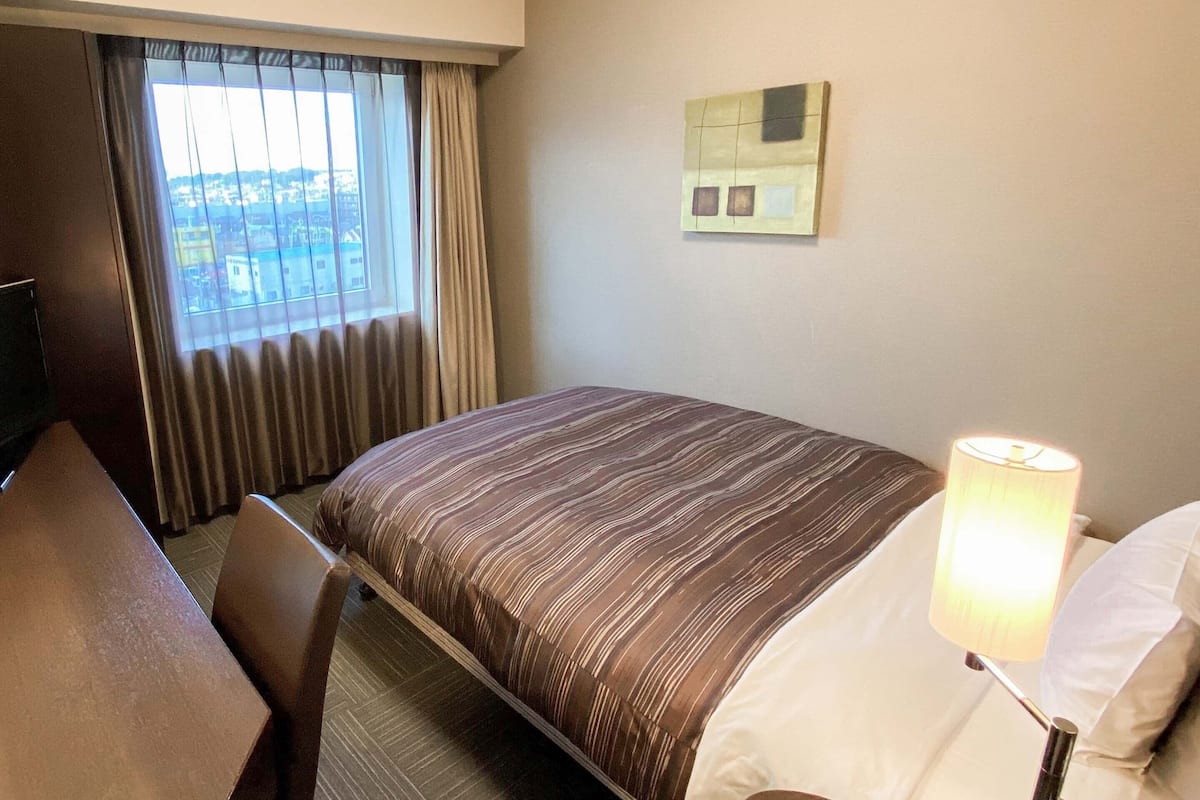 Ein Hotelzimmer mit Bett, Schreibtisch, Lampe und einem Fenster mit Vorhängen.