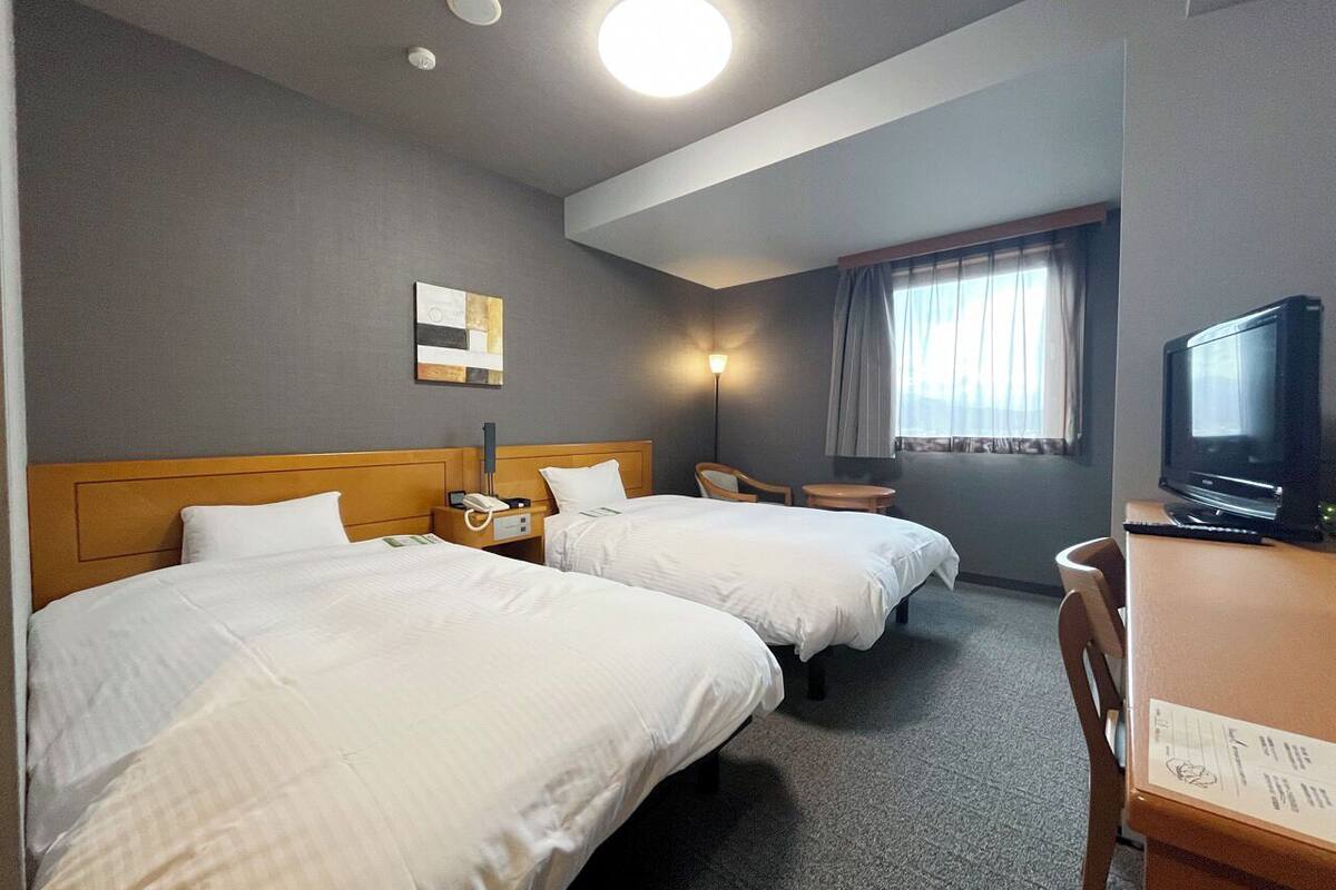 Una habitación de hotel con dos camas, un escritorio con televisor, una lámpara y una ventana con cortinas.