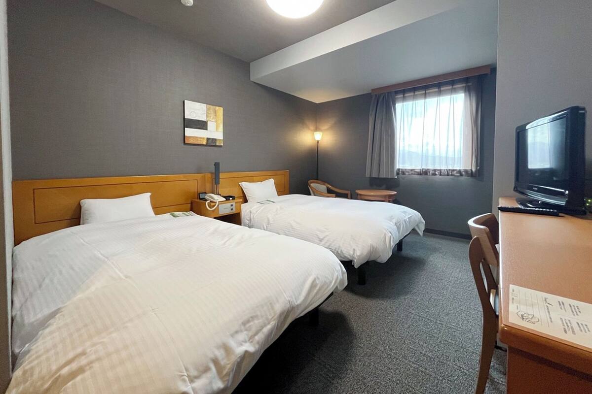 Habitación de hotel con dos camas, un escritorio con televisor, una silla y una ventana con cortinas.