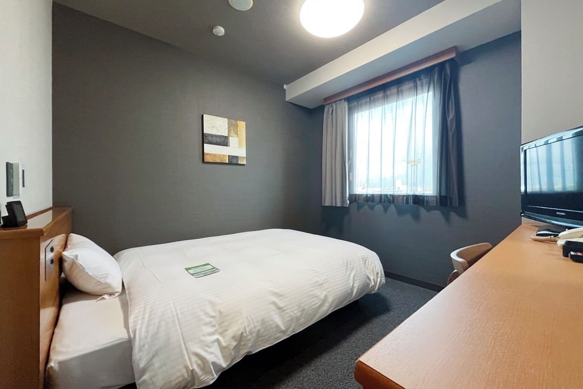Habitación de hotel con una cama individual, escritorio con computadora, televisor de pantalla plana, ventana con cortinas y un pequeño cuadro en la pared.
