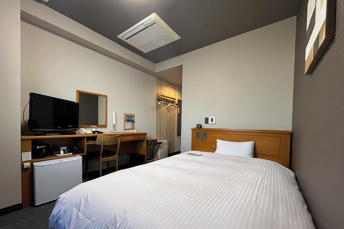 Habitación de hotel con cama, escritorio con computadora, televisor, mini refrigerador y espejo.