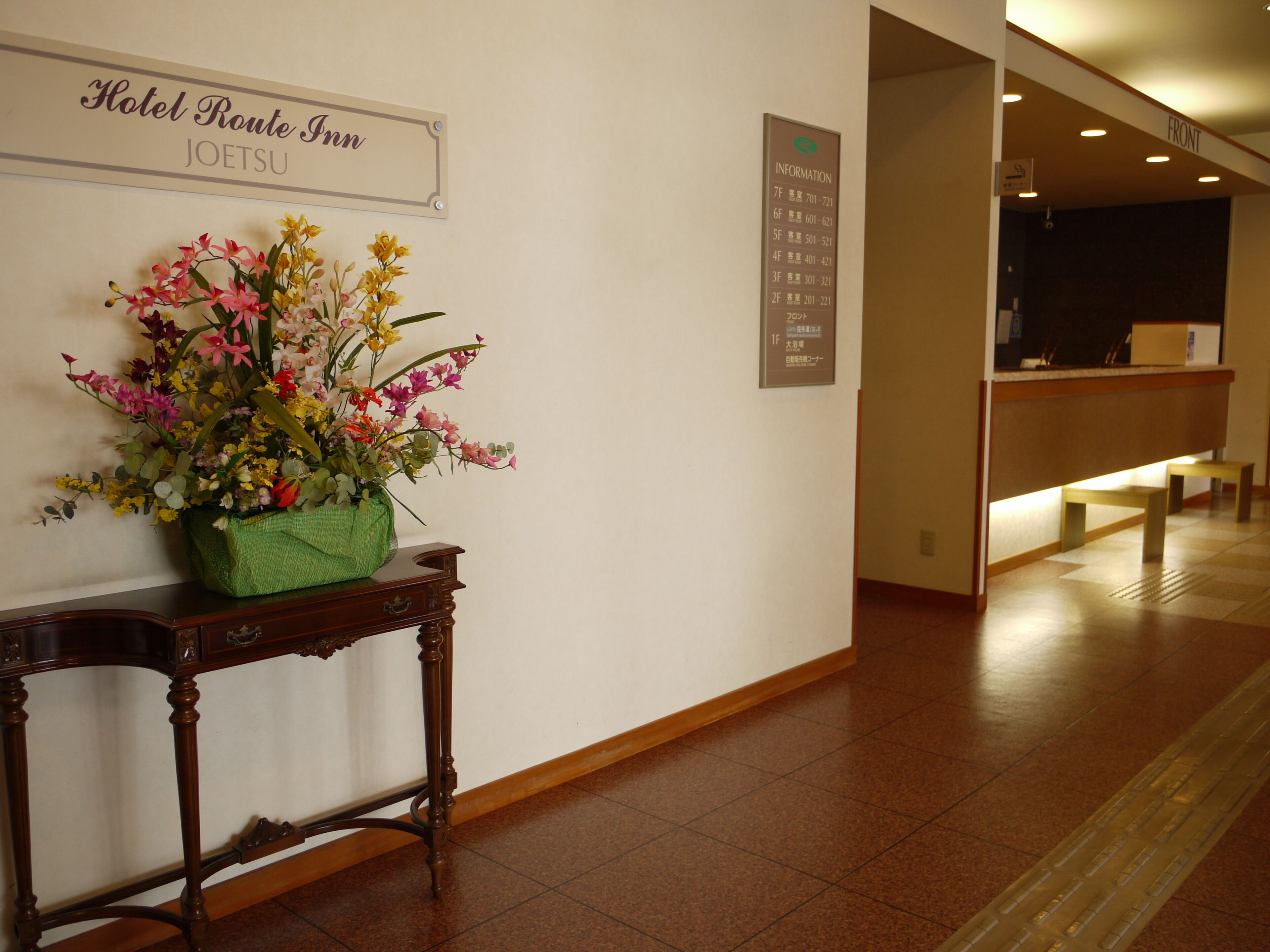Zona de recepción con un cartel que indica el nombre del hotel y detalles de las habitaciones. Un arreglo floral adorna una consola de madera.