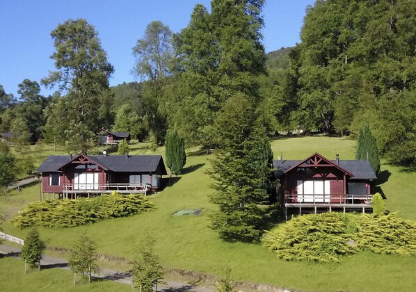 A cluster of cabins with a grassy hill and dense trees in the background. - Mirador Los Volcanes Lodge & Boutique (Pucón)