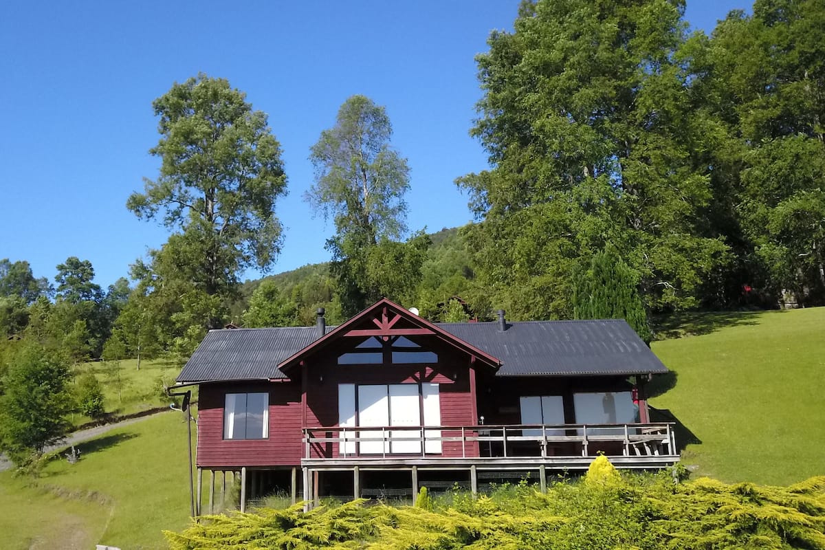 Une maison en bois rouge, dotée d’un balcon, entourée d’une végétation luxuriante et d’arbres.