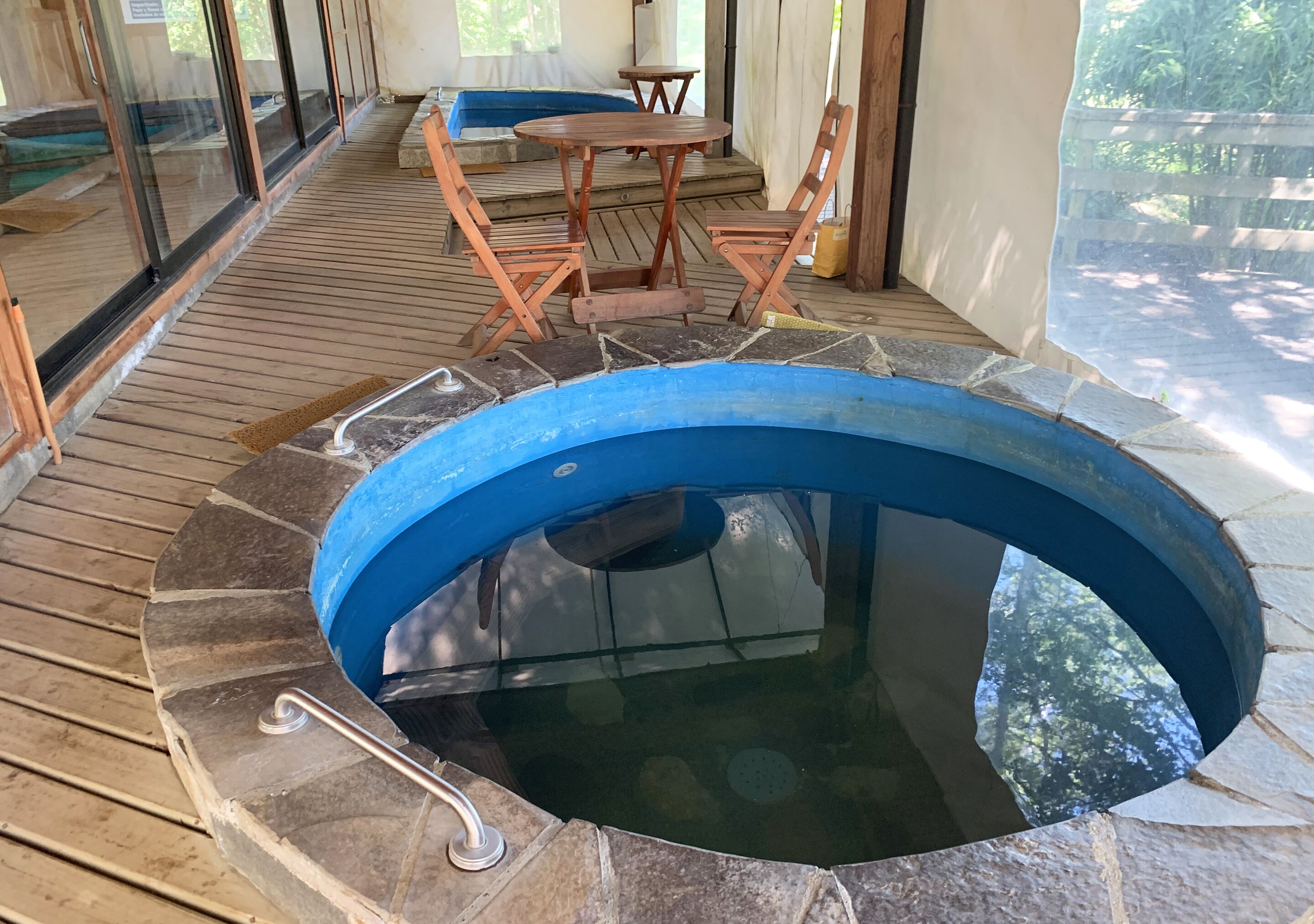 Un jacuzzi con un borde de piedra, muelle de madera y una mesa con sillas al fondo.