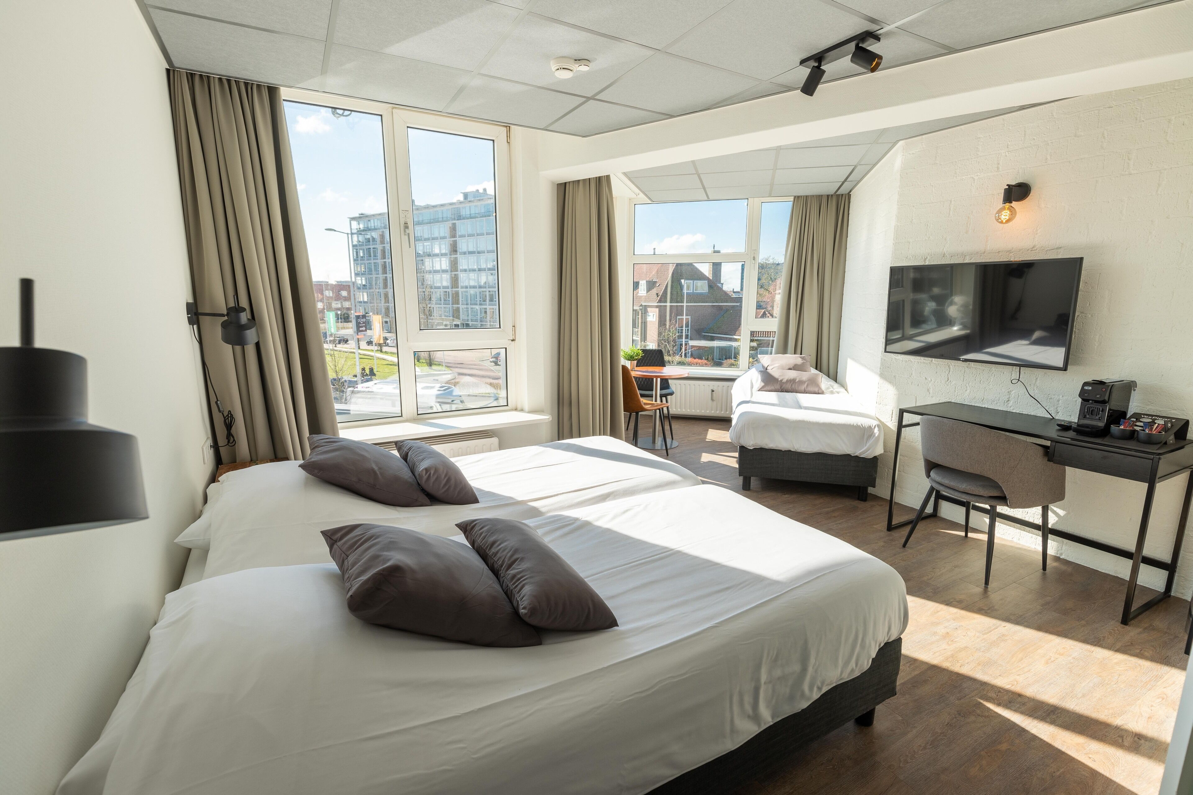 Habitación de hotel con dos camas, un escritorio, una silla, un televisor y ventanas amplias con vistas a la ciudad.