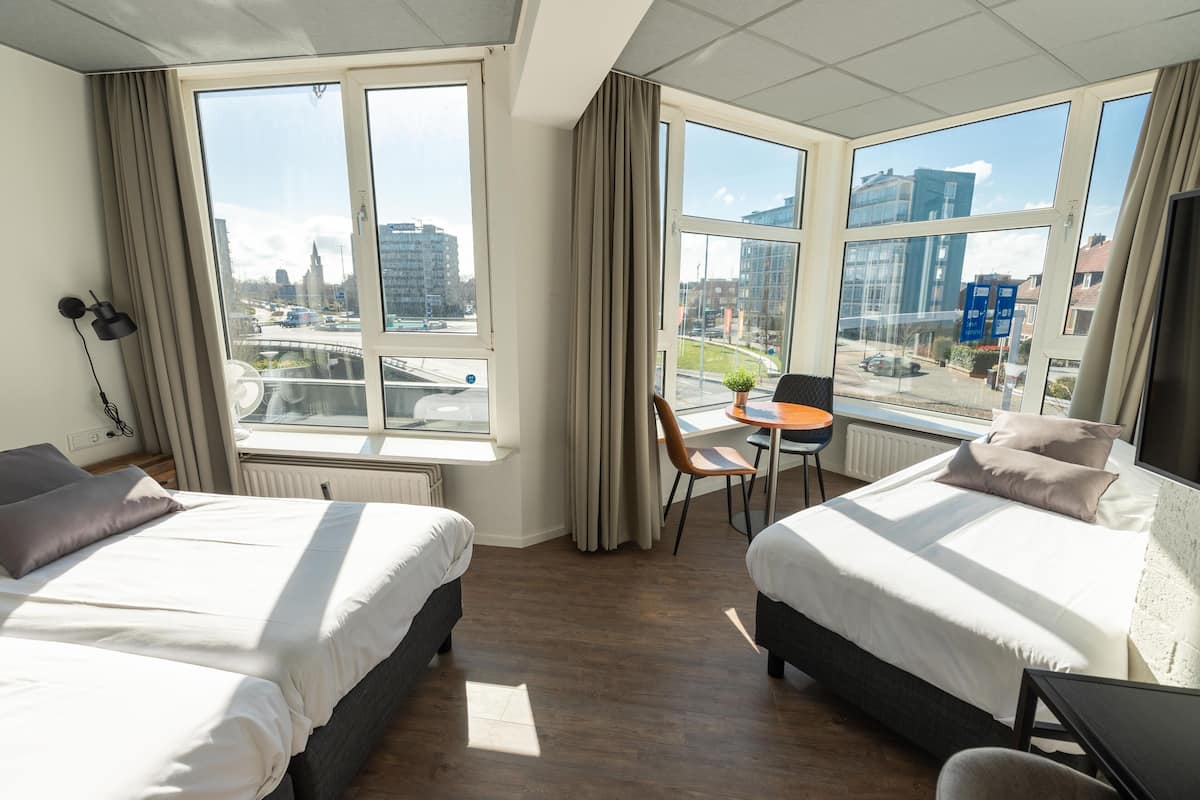 Een hotelkamer met twee bedden, een klein tafeltje en een stoel. Grote ramen bieden uitzicht op de stad.