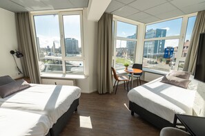 Een hotelkamer met twee bedden, een klein tafeltje en een stoel. Grote ramen bieden uitzicht op de stad.