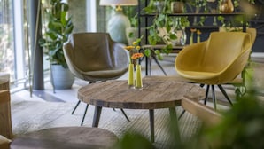 Una mesa redonda de madera con dos sillas, un jarrón con flores y una estantería con plantas en una habitación bien iluminada.
