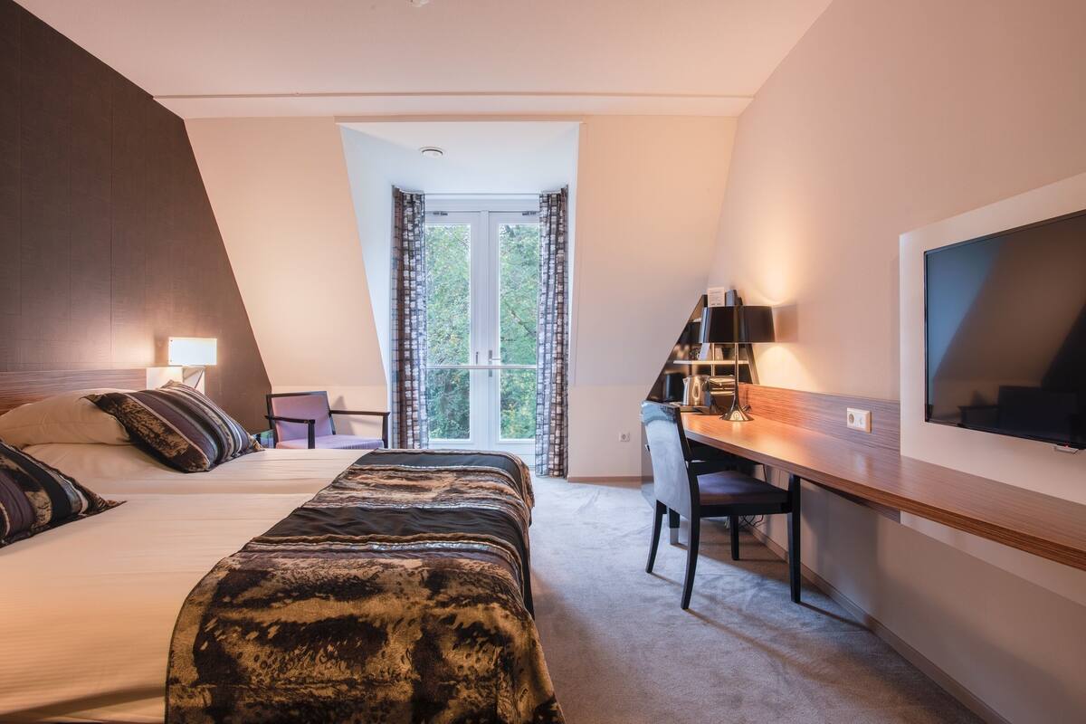 Un dormitorio con cama, escritorio, silla, televisor y ventana con cortinas.