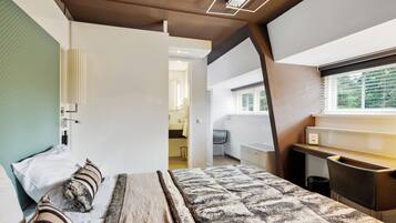 Un dormitorio con cama, almohadas, una mesita de noche, lámpara, ventana con persianas y vista al baño.