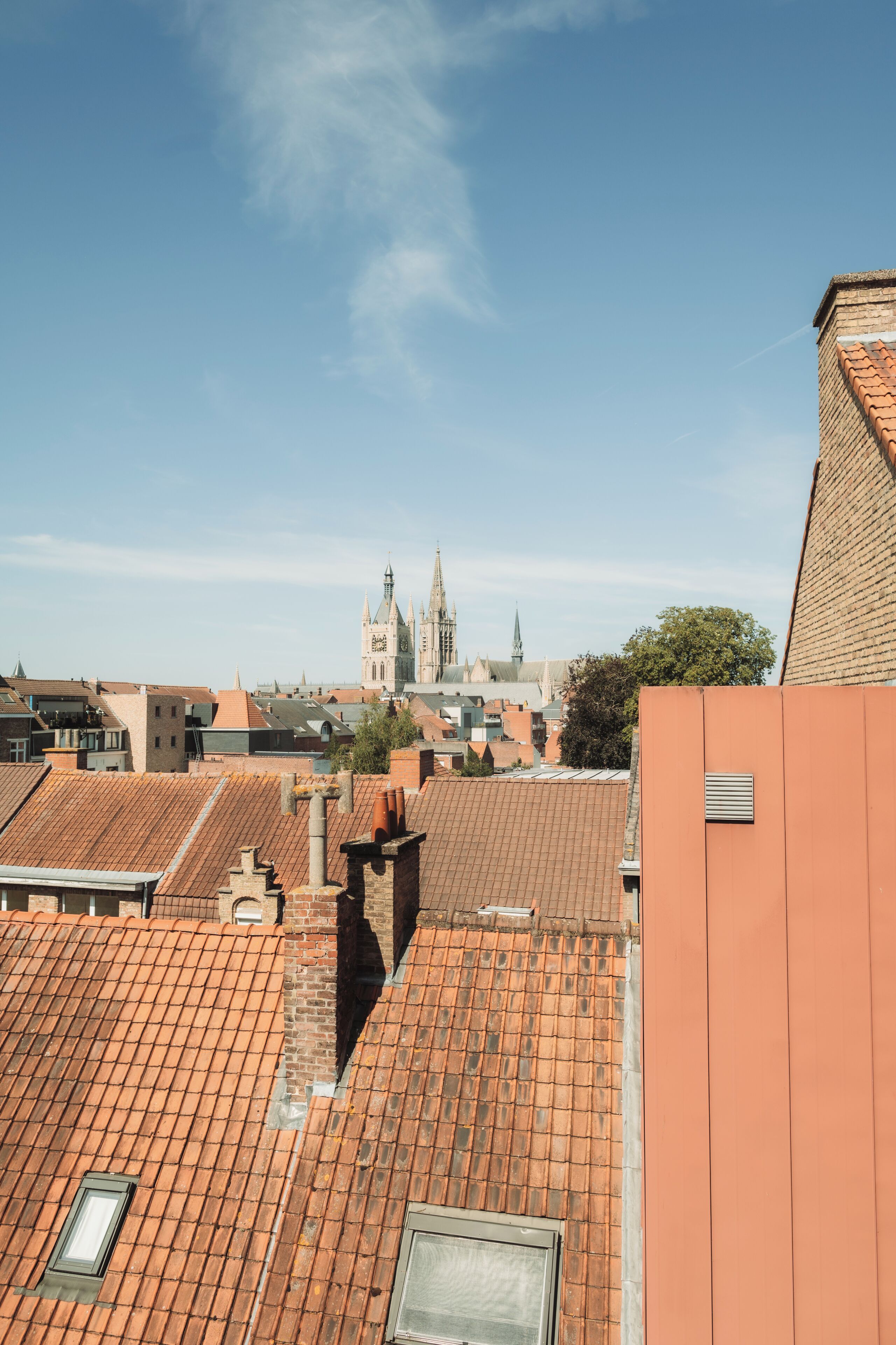 A view of rooftops with terracotta tiles, a distant church spire, and a clear blue sky.