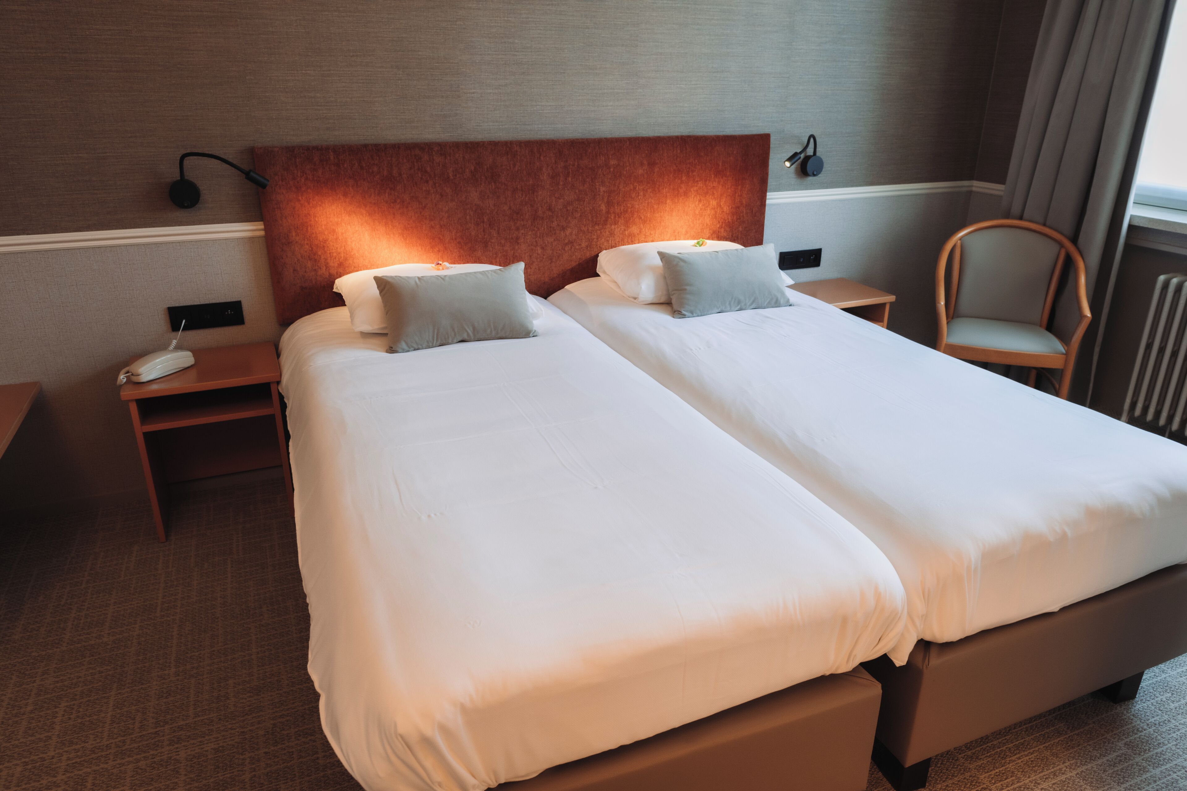 A hotel room with two beds, a nightstand with a phone, a chair, and a wall-mounted lamp.
