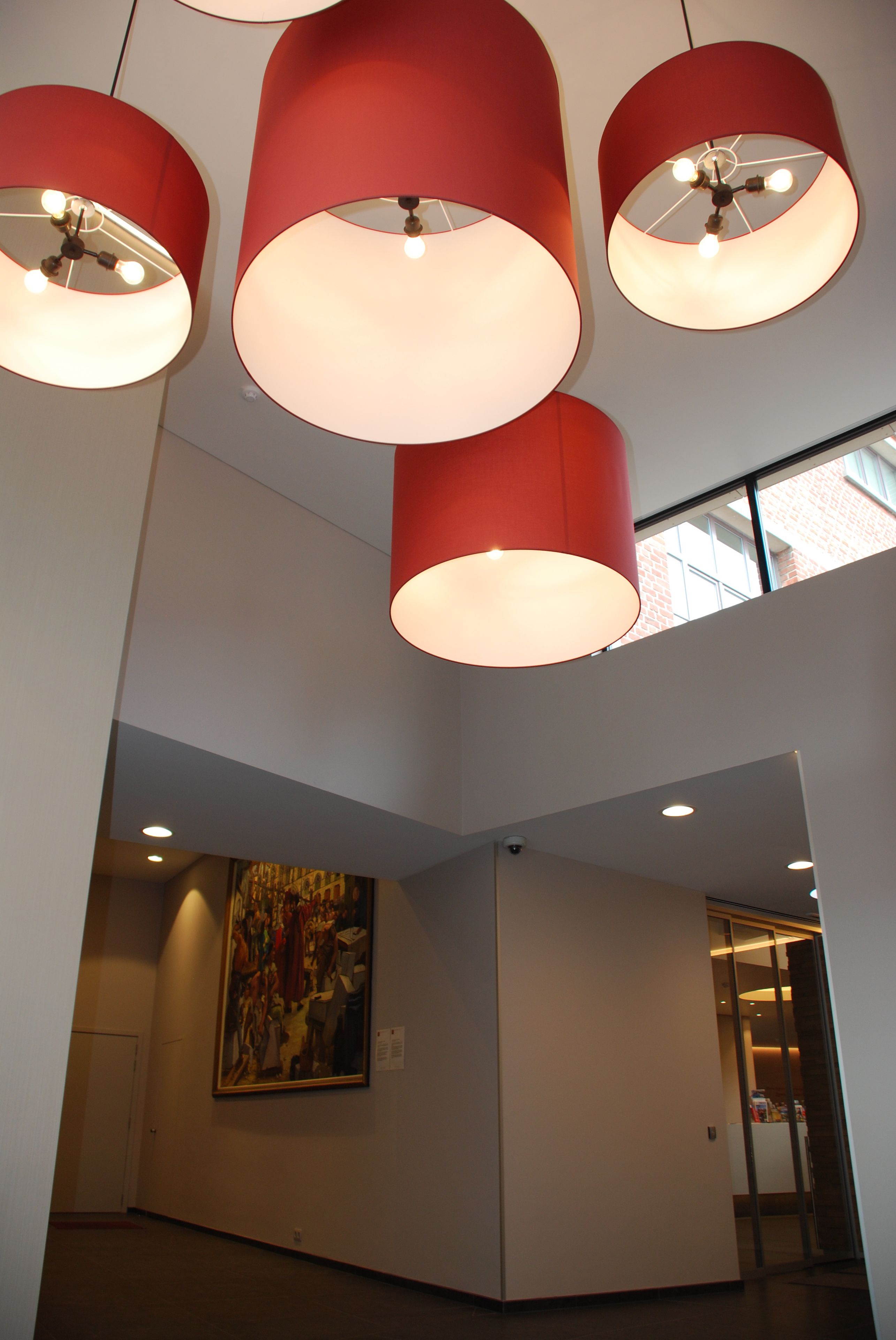 Um interior moderno com uma pintura grande na parede, uma luminária cilíndrica vermelha e uma porta de vidro que dá para outro cômodo.