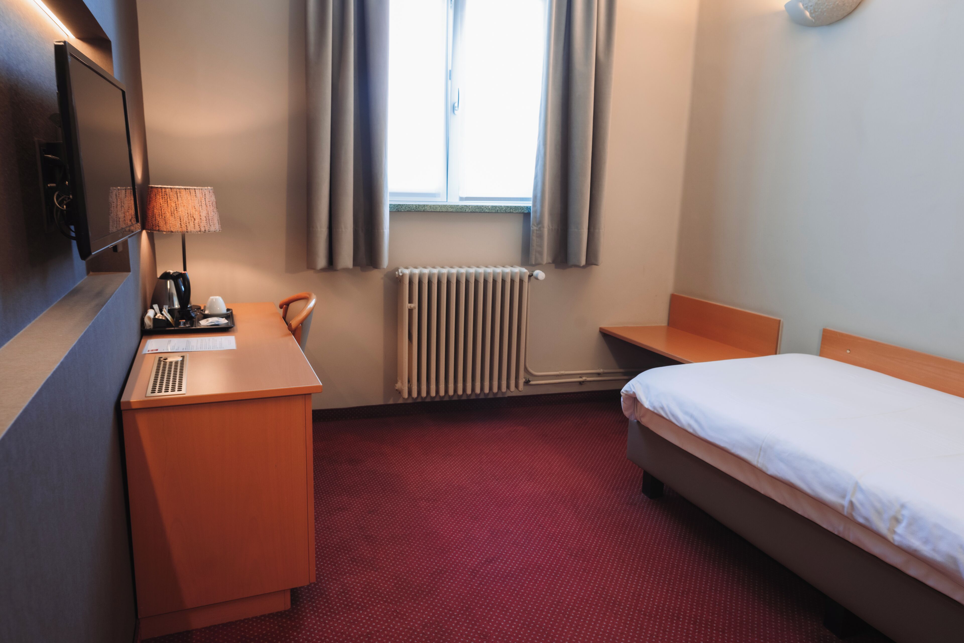 A hotel room with a bed, a desk, a chair, a TV, a lamp, a window with curtains, and a radiator.