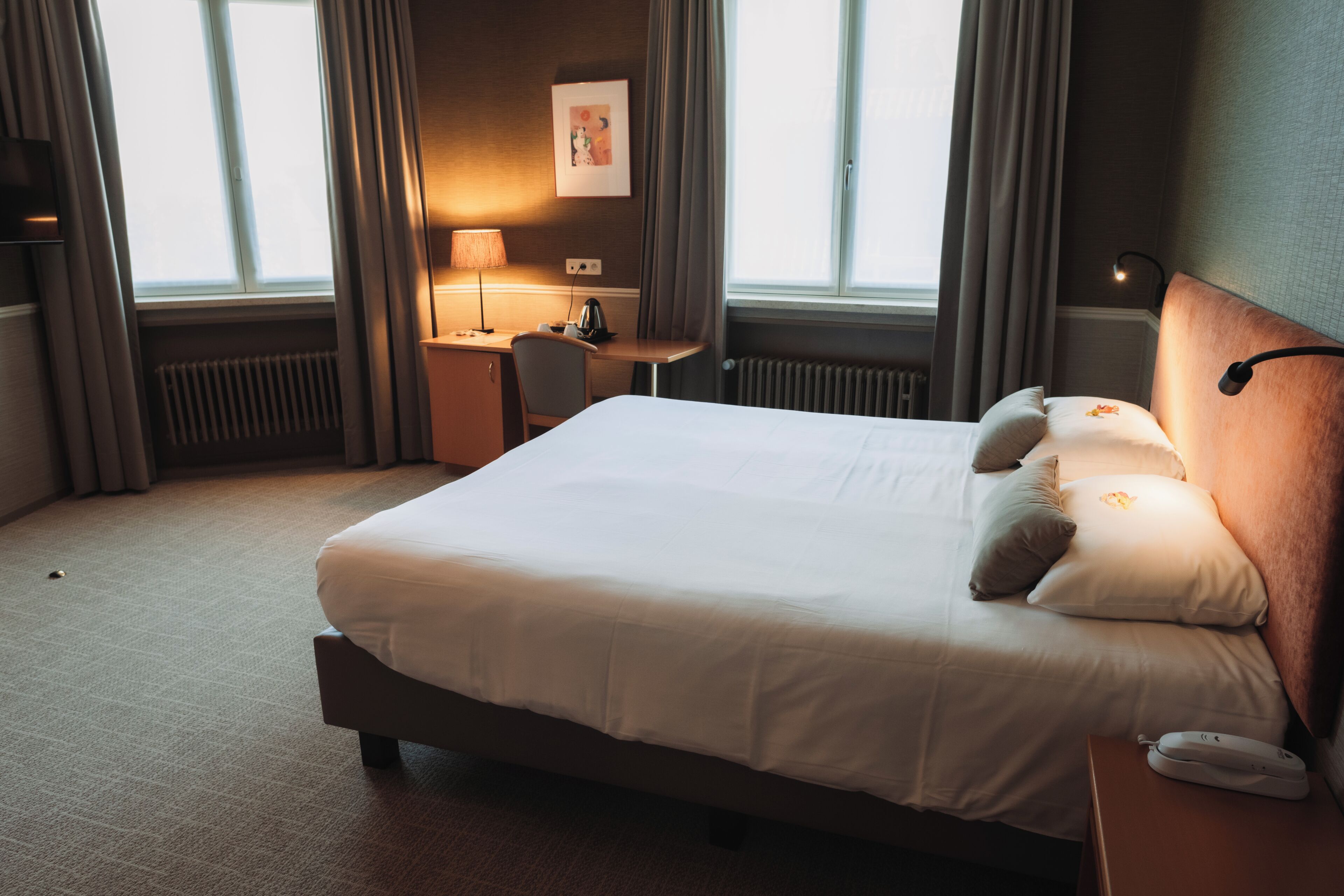 Une chambre d’hôtel avec un lit, un bureau, une chaise, une lampe et une fenêtre.