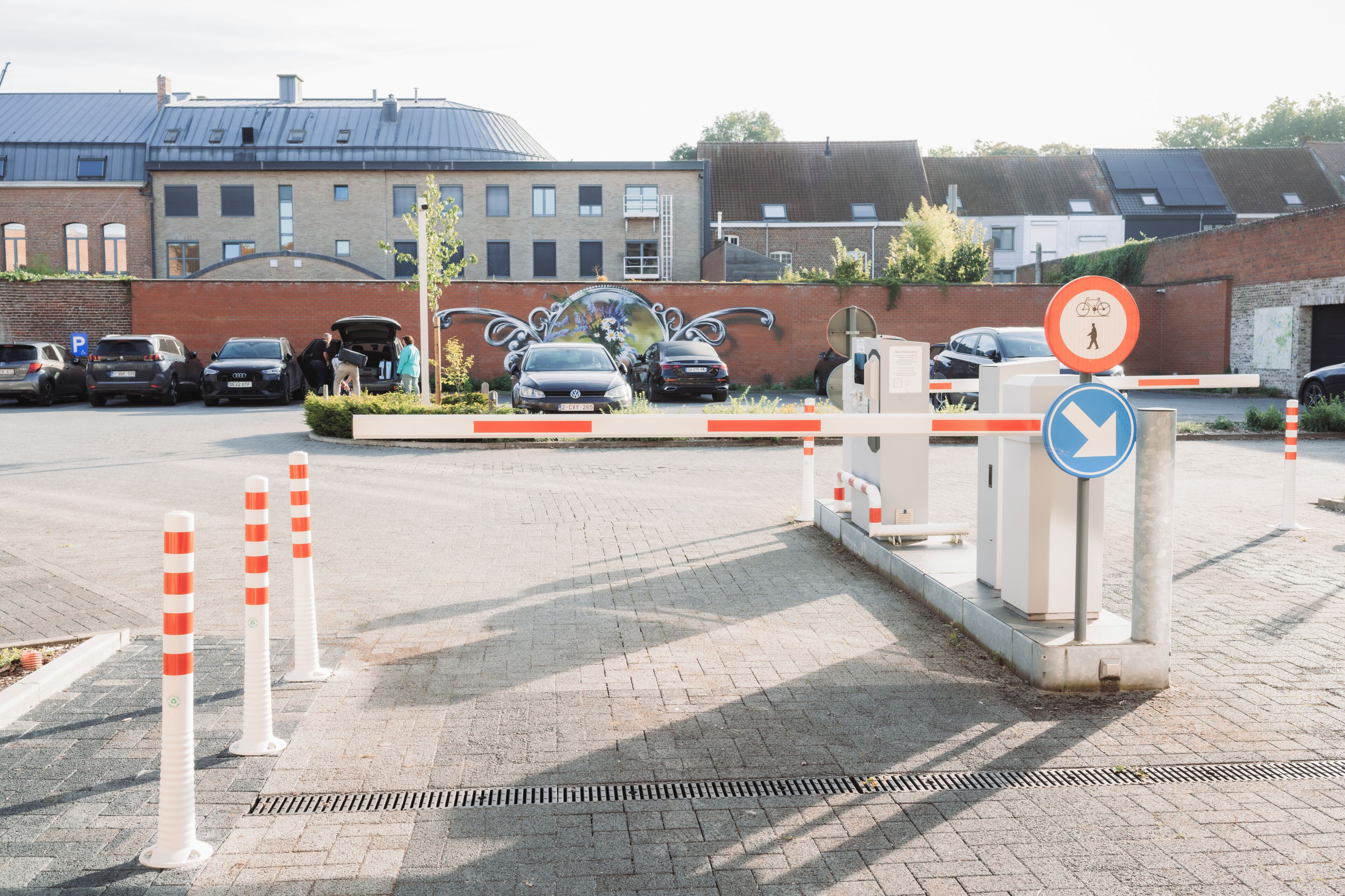 A gated parking area with a blue directional sign, a brick building, and a mural on the wall.