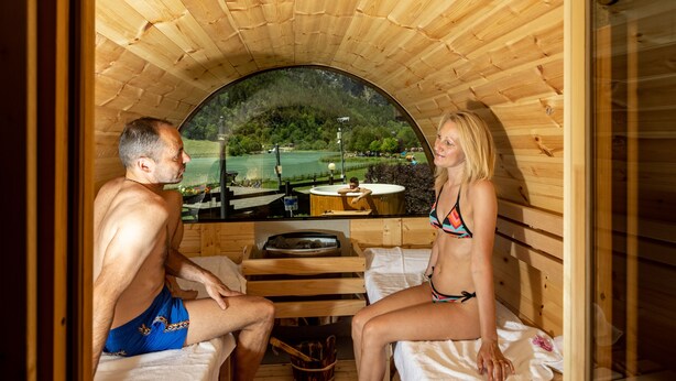 A wooden sauna with a curved roof, a wooden bench, and a view of a lake and trees. - Hotel Laghetto (Brusson)