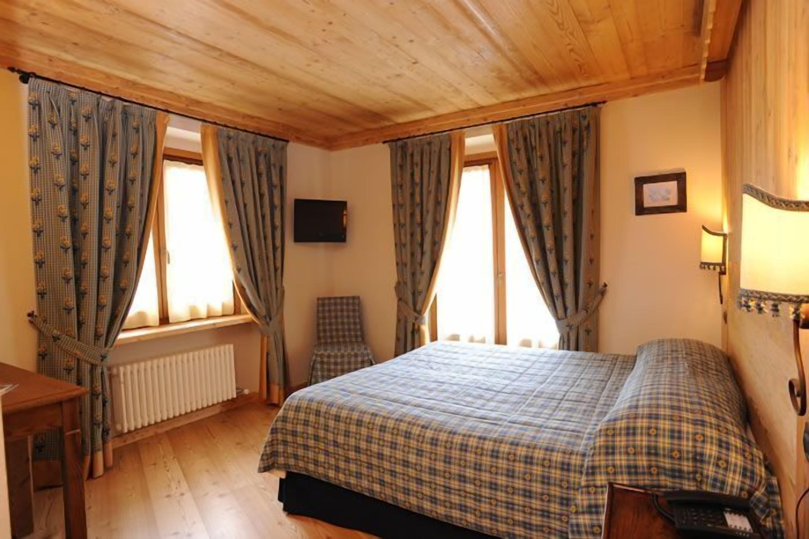 A bedroom with a wooden ceiling, a bed with a plaid bedspread, two windows with curtains, a chair, a radiator, and a wall-mounted lamp.