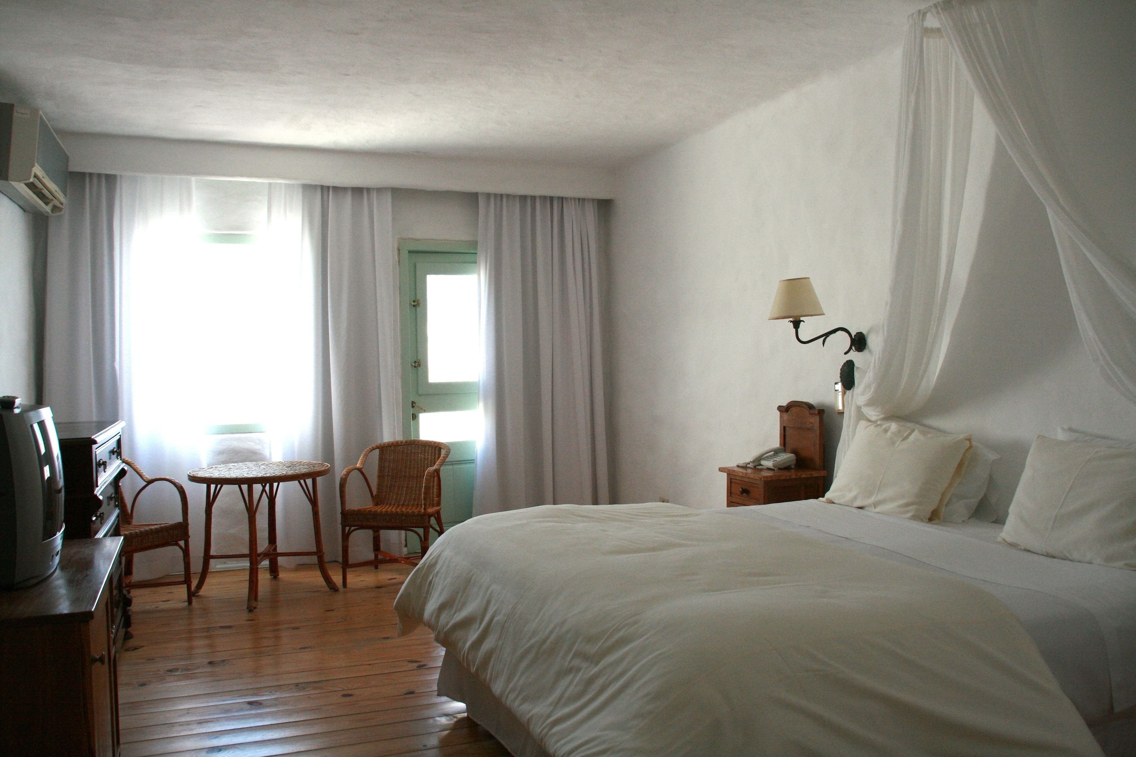 A hotel room with a large bed, a small table, two wicker chairs, a nightstand with a lamp, and a window with curtains.