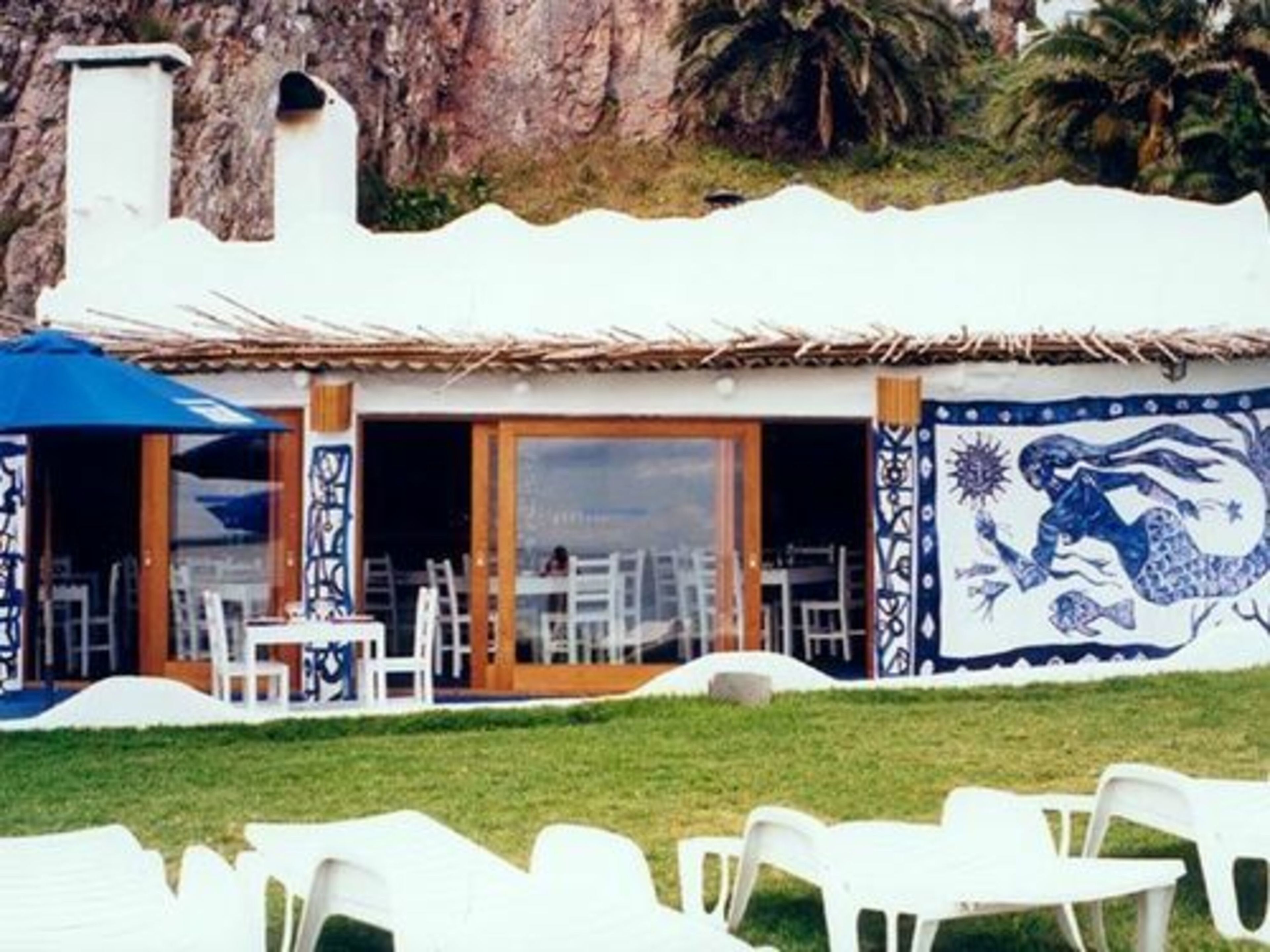 A restaurant with outdoor seating, white chairs, and a blue umbrella.