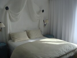 Una cama con sábanas blancas, una mesita de noche, una lámpara y tela decorativa en la pared.