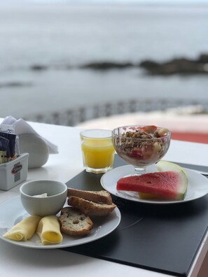 Bandeja de café da manhã com pão, uma tigela de iogurte, um copo de suco de laranja e uma fatia de melancia sobre uma mesa perto de uma janela com vista para o mar.