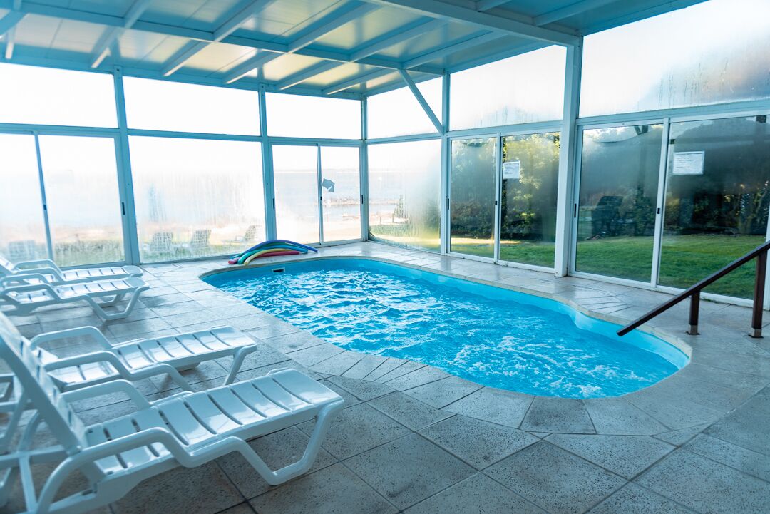 Una piscina interior con paredes de vidrio transparente, sillas de descanso y vistas del entorno.
