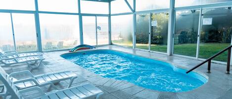 Una piscina interior con paredes de vidrio transparente, sillas de descanso y vistas del entorno.