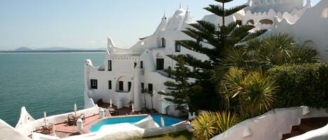A white building with a pool and stairs leading up to it, overlooking a body of water.