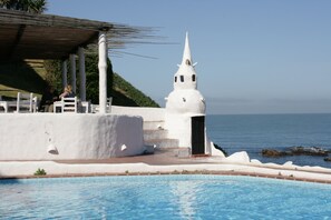 Edifício branco com telhado pontiagudo próximo a uma piscina, com vista para o mar.