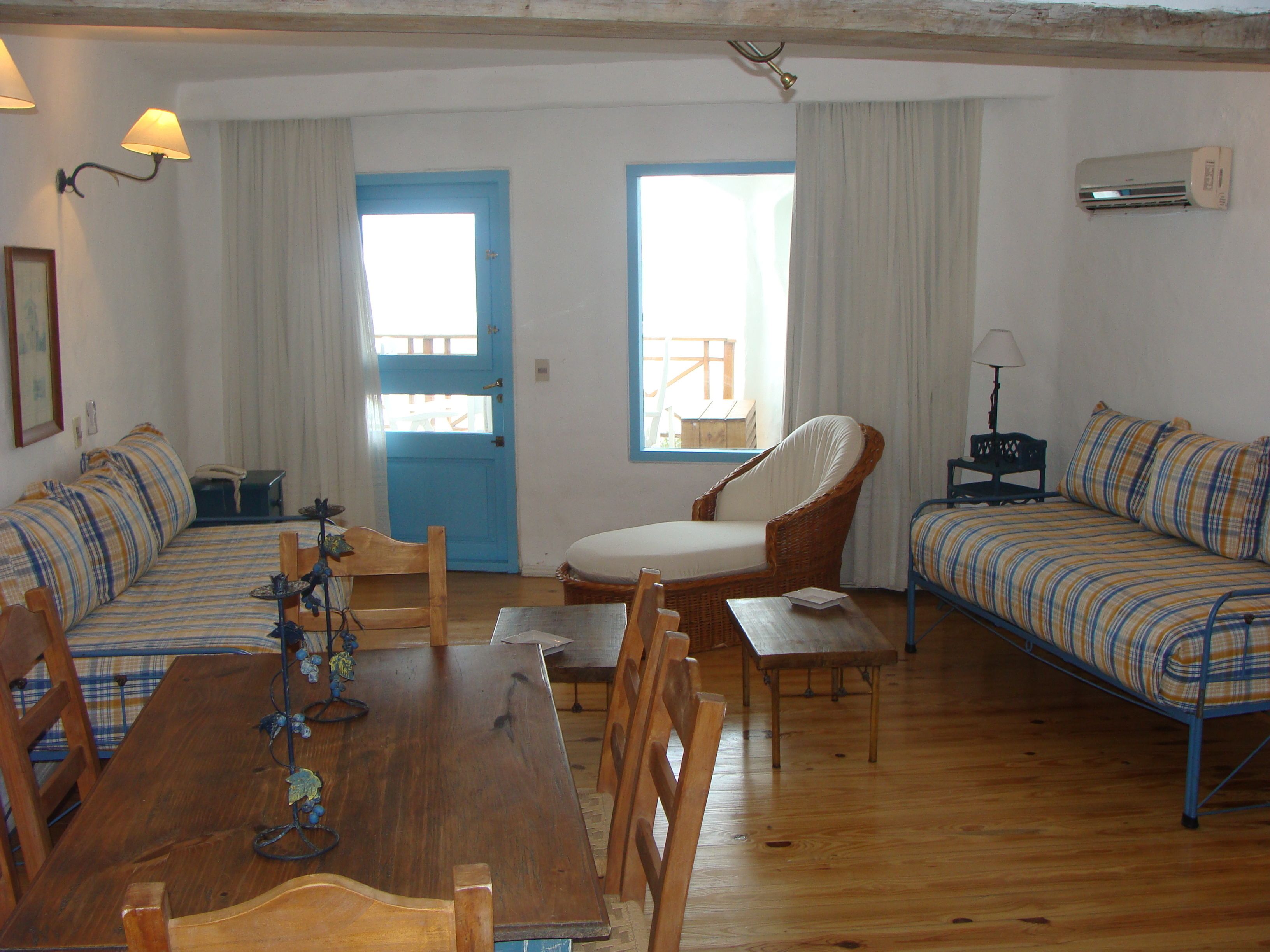 A room with a wooden dining table, two sofas with plaid upholstery, a wicker chair, and a small table with a lamp.