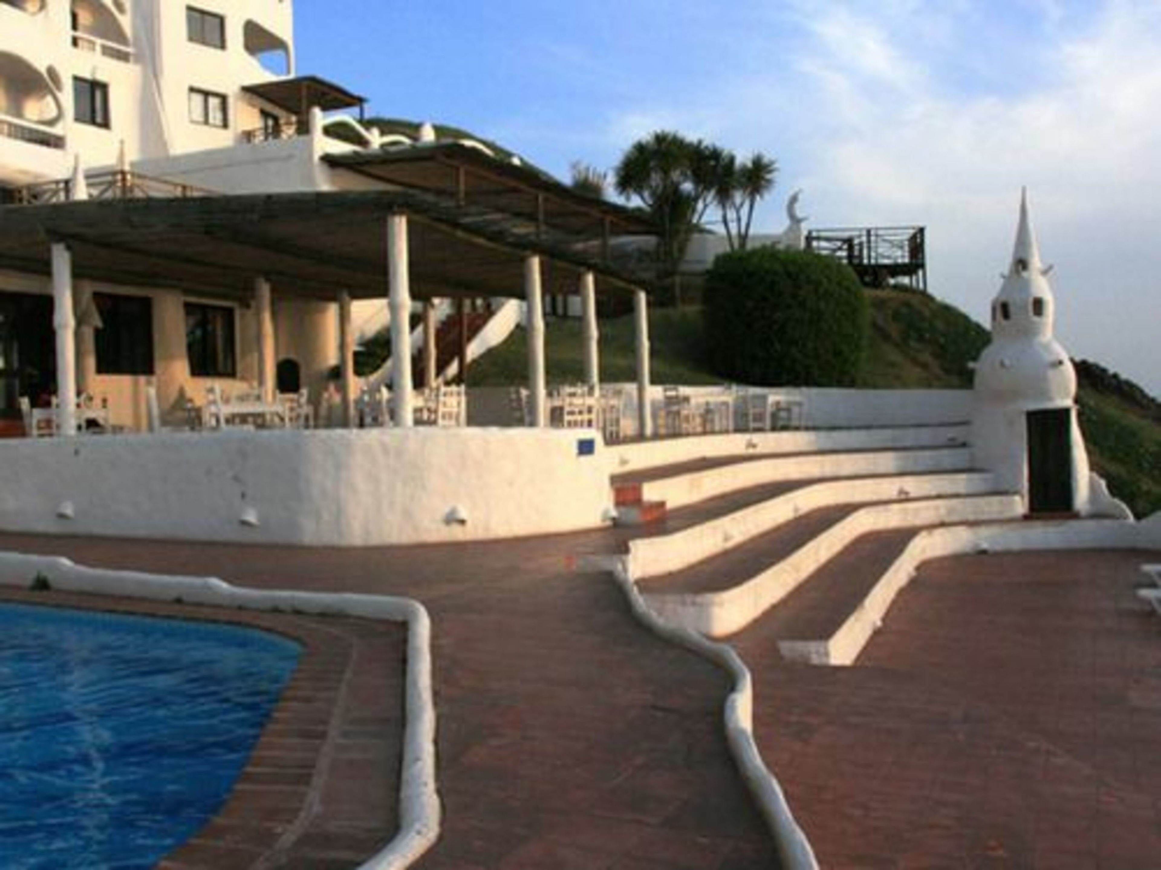 A pool area with a curved edge, steps leading up to a white structure, and a building with balconies in the background.