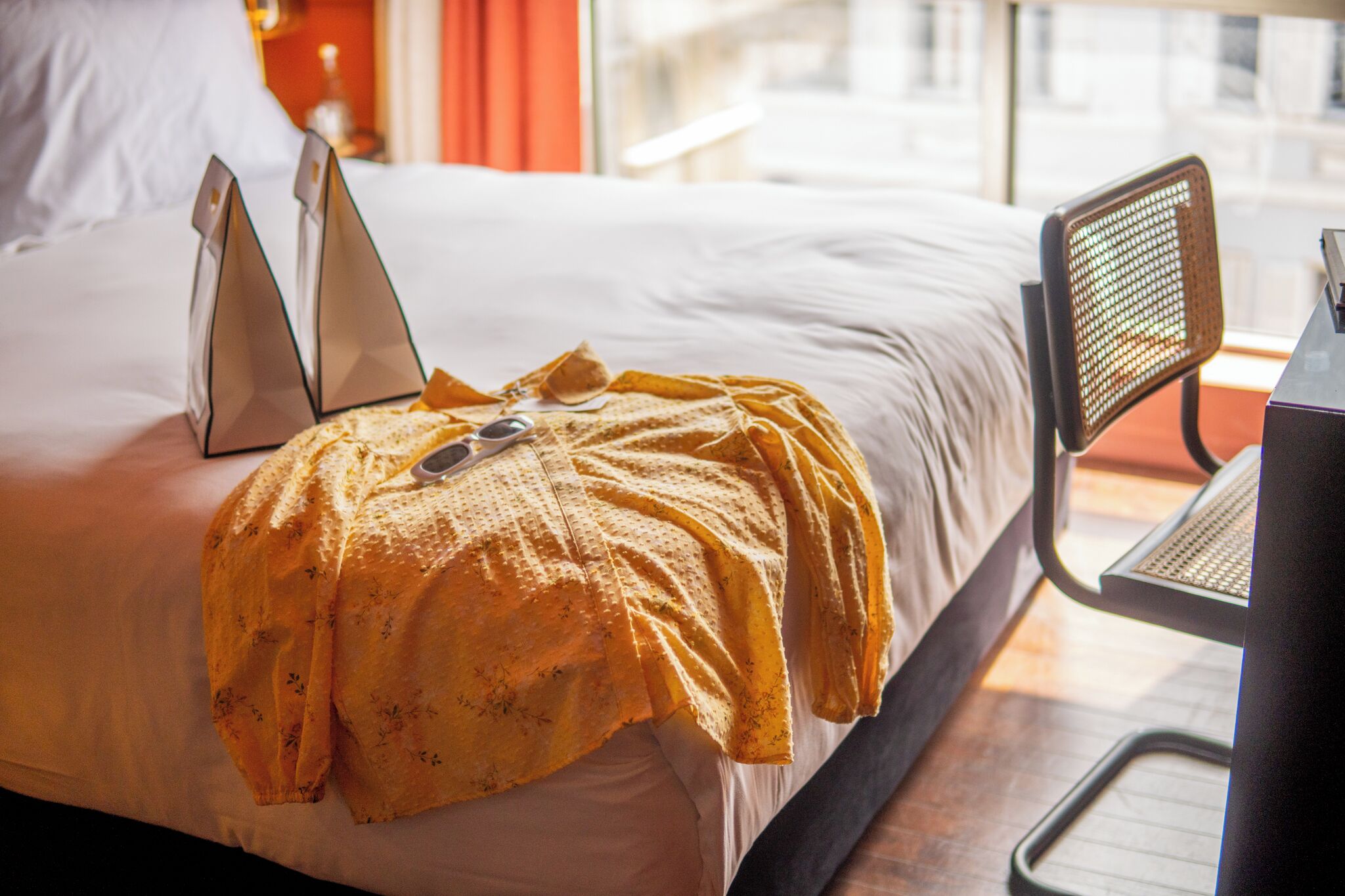 Una cama impecable con una computadora portátil y gafas de sol encima, una silla y vista de la ciudad a través de una ventana.