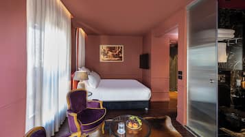 Habitación de hotel con cama, un sillón morado, una mesita con fruta y un cuadro en la pared.